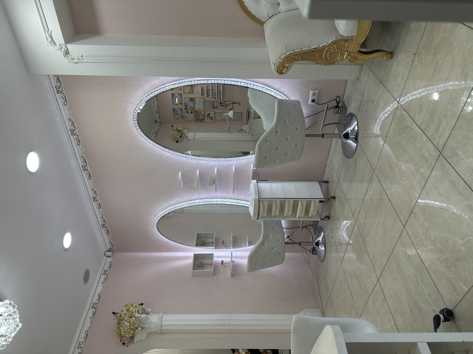 The image shows the interior of what appears to be a beauty salon, possibly in Sofia, Bulgaria, given the European-style architectural details. The room has light pink walls with ornate white molding and two oval-shaped mirrors with LED lighting around the edges. Each mirror reflects a white, tufted salon chair. A rolling white cabinet is positioned between the two chairs. The floor is tiled in a light, neutral color with a glossy finish. There is a white column adorned with flowers in the lower left corner, and a glimpse of an ornate chair is visible in the upper right. The lighting appears to be a combination of natural light and artificial light fixtures, including recessed ceiling lights. The overall impression is a clean, stylish, and well-lit space.