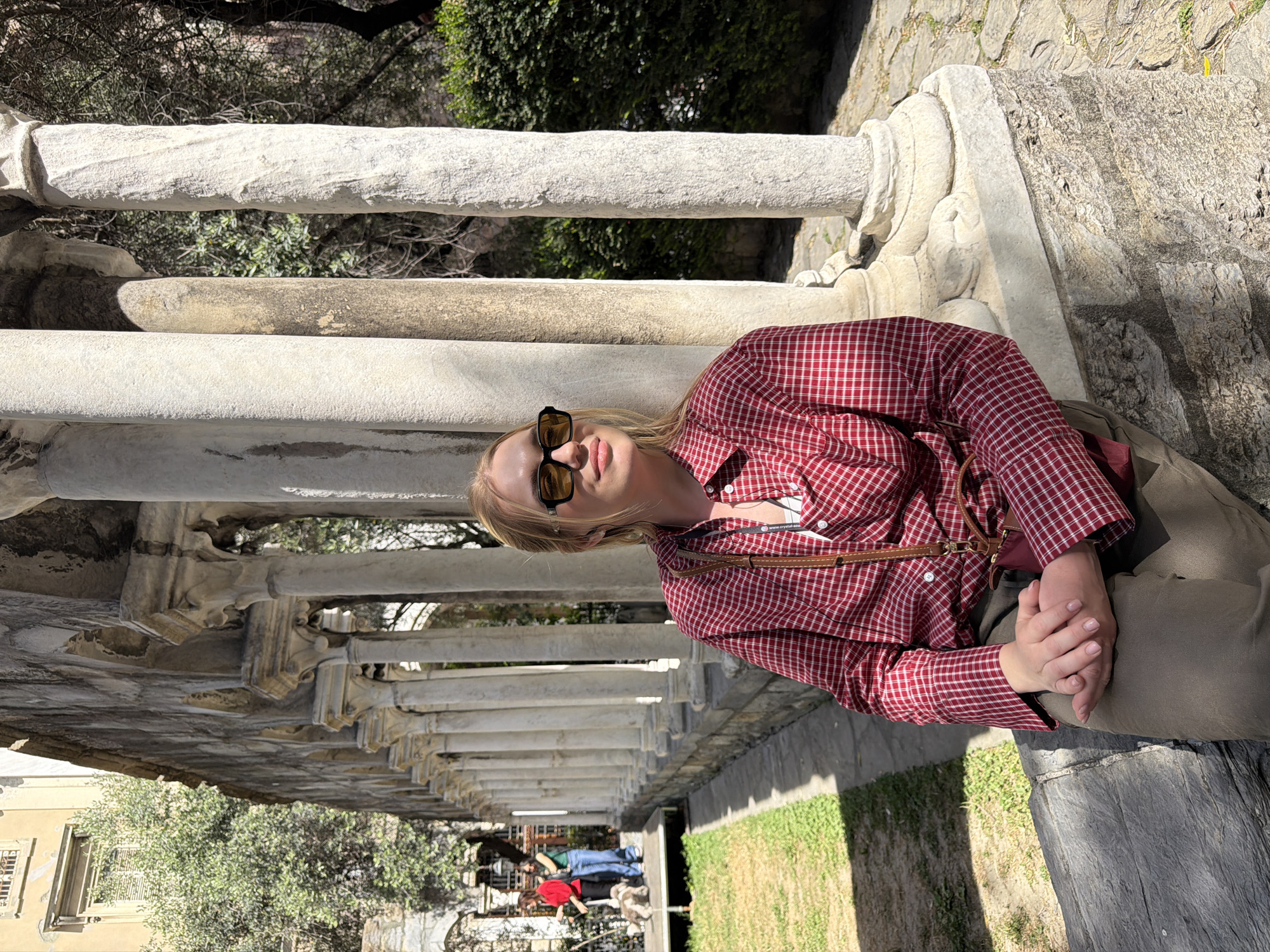 A medium, portrait-oriented shot captures a woman wearing sunglasses and a red plaid shirt, positioned against a row of stone columns. She sits with her hands clasped in her lap. The columns form a covered walkway with glimpses of trees and buildings in the background. People are seen in the distance. The location is Genoa, Italy.