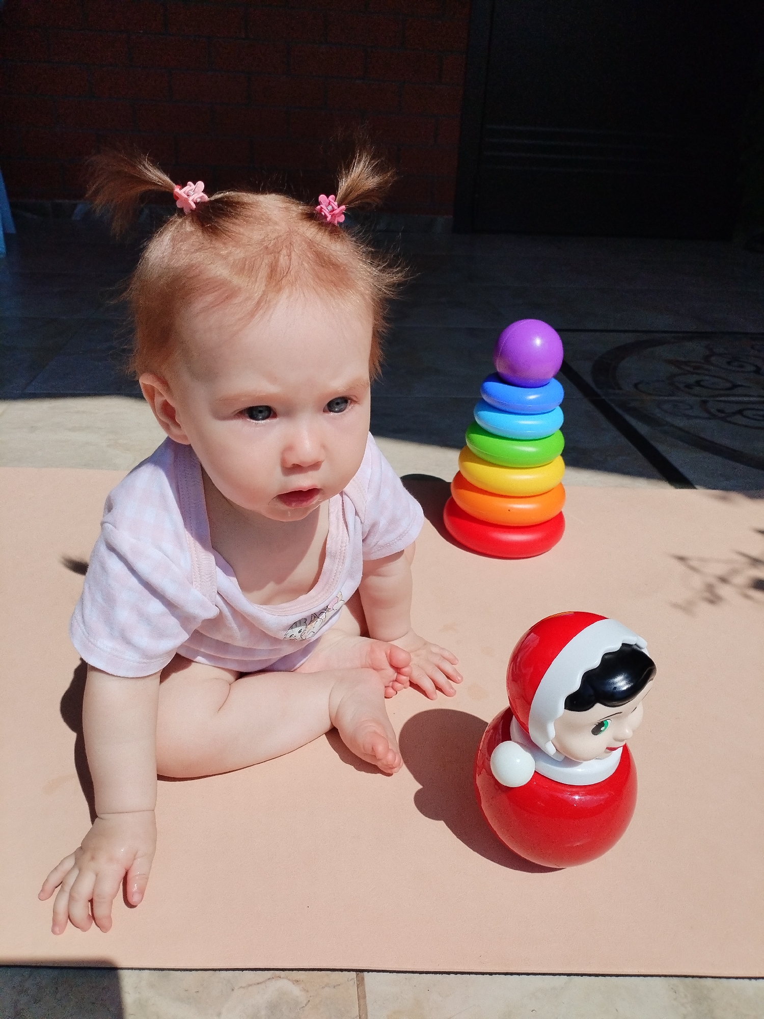 A fair-skinned baby with red hair styled into two pigtails sits on a light pink mat outdoors. The baby is wearing a light pink and white checkered onesie and is looking to the right of the frame. Next to the baby is a red tumbler toy and a stacked ring toy with colorful rings. The background shows a brick wall, a dark door, and a tiled floor. The scene is lit by harsh sunlight, casting shadows on the mat and floor.