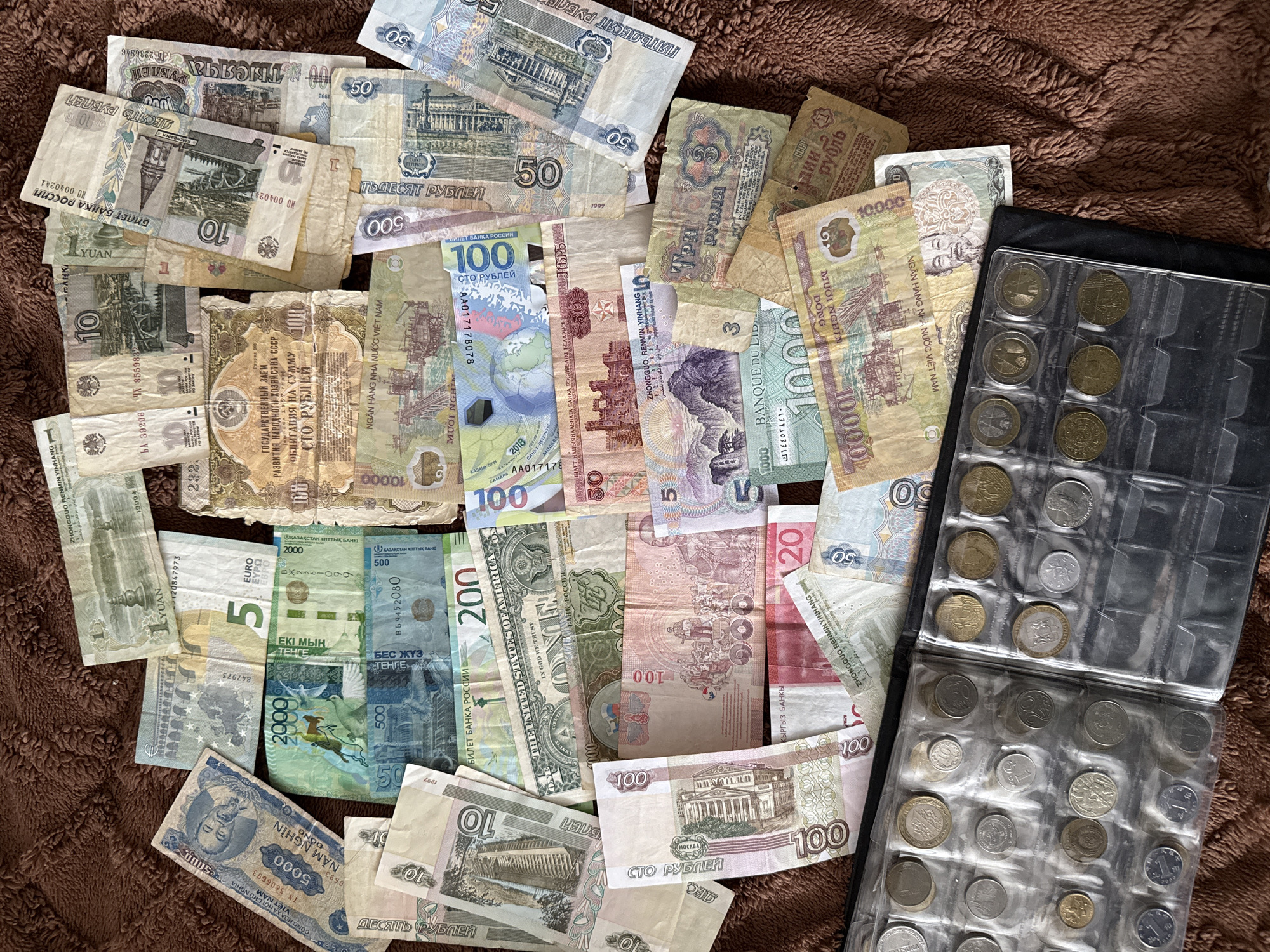 The image shows a collection of world currencies and a coin collection book on a brown, textured surface, possibly fabric. Banknotes from Russia, Vietnam, China, Kazakhstan, Belarus, Kyrgyzstan, the USA, and the EU are scattered across the surface. The banknotes vary in denomination, from 1 to 100,000. The coin collection book has multiple slots, each holding a coin of varying size, color, and design. The scene appears to be a collection rather than a transaction or event, and there is no visible indication of the city of Mirny, Russia. No people or actions are visible.