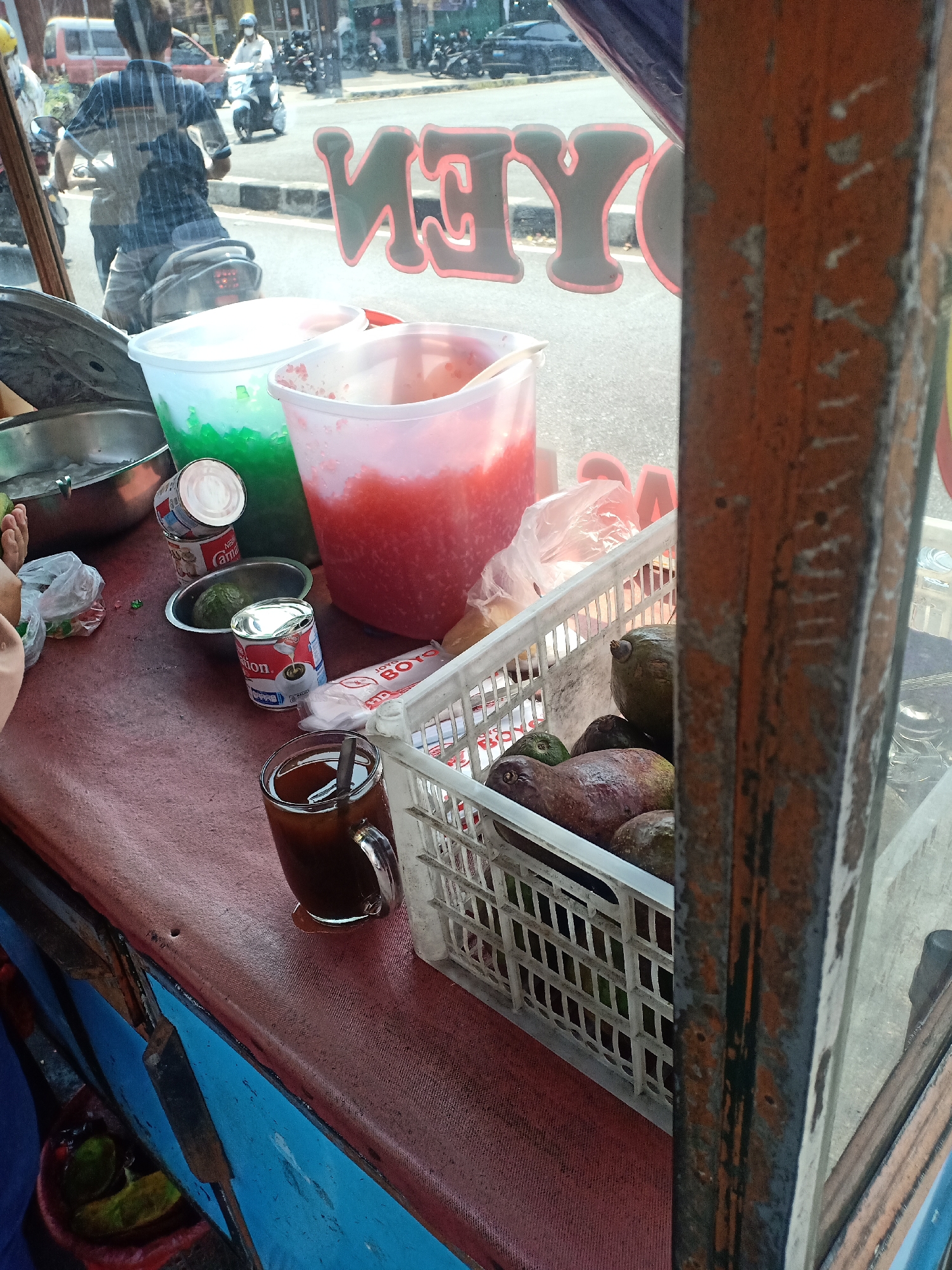 The image shows a street food stall in Semarang, Indonesia, likely operating on a sunny day. In the foreground, a red-covered table displays various items for sale. To the left, a large metal bowl contains what appears to be ice. Next to it are two large plastic containers, one filled with green shaved ice and the other with red liquid, possibly a fruit-based drink. Two cans of condensed milk, one labeled "Carnation" and another "Nona" (likely sweetened condensed milk), are stacked on the table. A bowl with an avocado sits in front of them. A glass filled with a dark beverage, likely coffee or iced tea with ice and a spoon, is positioned in the center of the table, with a condensation ring forming on the surface. To the right, a white plastic basket overflows with avocados. A package labeled "BOTO" is also visible near the basket. In the background, through the glass of the stall, traffic and people on motorcycles are visible on a street, suggesting a bustling urban environment. The overall impression is of a typical roadside eatery preparing to serve customers. The stall itself appears to be made of metal and wood, with a rustic and well-used appearance. The presence of avocados, shaved ice, and condensed milk suggests that the stall sells beverages and possibly desserts.