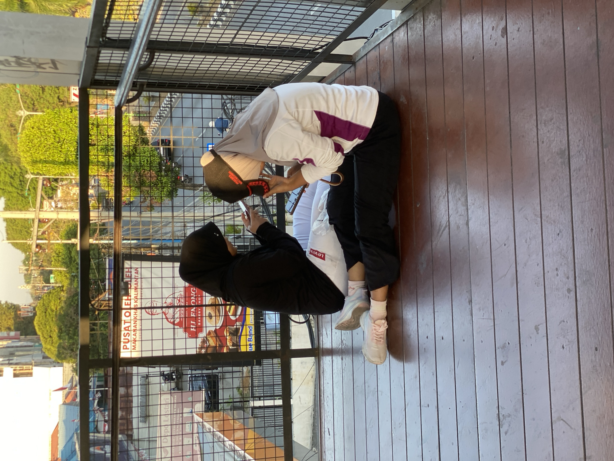 The image shows two young women sitting on a wooden deck, facing each other and looking at their phones. They are both wearing hijabs and casual clothing. The woman on the left is wearing a black hijab and a black long-sleeved shirt with white pants. The woman on the right is wearing a grey hijab, a white and purple long-sleeved shirt, and black pants. Both are wearing sneakers. The scene appears to be on an elevated walkway or balcony overlooking a street in Banjarbaru, Indonesia. Behind them, a fence separates them from the street, where traffic and lush green trees are visible. A sign in the background reads "Pusat Oleh Oleh Makanan Khas Kalimantan" and "HJ EN ONG BAKERY SHOP," indicating a local souvenir and food shop. The lighting suggests it is late afternoon or early evening.