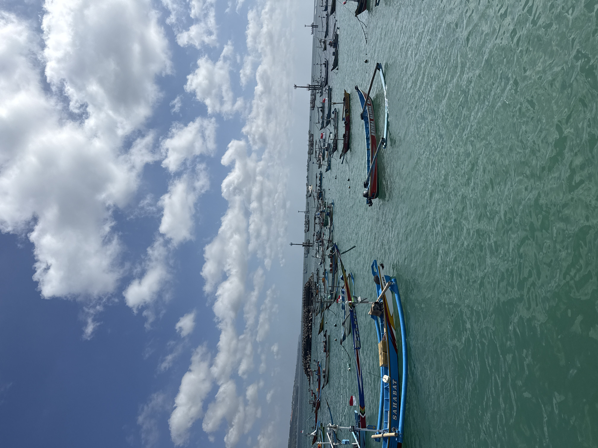 The image shows a harbor with many traditional Indonesian fishing boats, called "Jukung", on the water. The boats are painted in bright colors such as blue, red, and yellow. The boat closest to the viewer has "SAHABAT" written on its side. The water is a clear turquoise color. A line of buildings on the horizon may be Kuta in Indonesia. Above the harbor, the sky is filled with puffy white clouds against a blue sky.