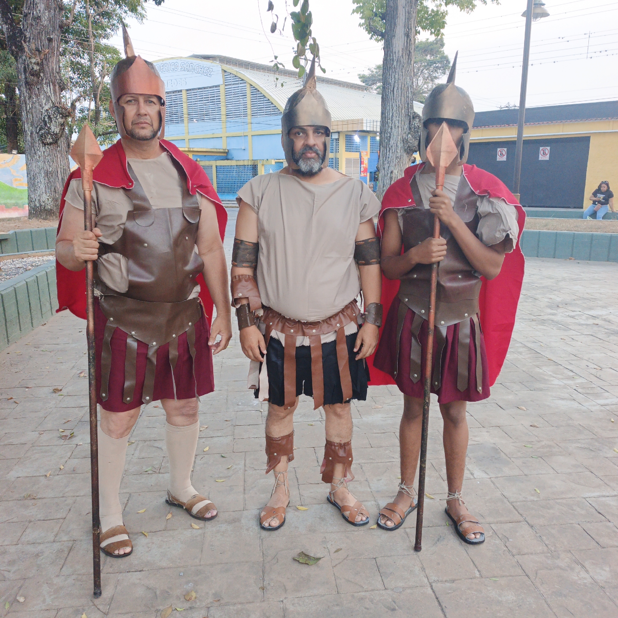 Three individuals in ancient soldier costumes are standing upright on a paved outdoor surface in Tinaquillo, Venezuela, with the "Gimnasio Cubierto Federico Sanchez" building visible in the background.
The leftmost subject is an adult male wearing a copper-colored spiked helmet, a light tan tunic, a red cape, brown faux-leather body armor and skirt flaps over a maroon skirt, light-colored knee-high socks, and brown sandals. He holds a brown spear with a copper-colored blade in his right hand.
The central subject, an adult male with a prominent gray beard, wears a darker copper-colored spiked helmet, a light tan tunic, brown faux-leather armbands, a waist belt with skirt flaps over a dark blue/black skirt, brown faux-leather shin guards, and brown ankle-strapped sandals. His hands are at his sides.
The rightmost subject, a younger male, wears a darker copper-colored spiked helmet with a face visor, a light tan tunic with ruffled sleeves, a red cape, brown faux-leather body armor and skirt flaps over a maroon skirt, and brown ankle-strapped sandals. He holds a brown spear with a copper-colored blade in his left hand, partially obscuring his face.
All three individuals face the camera. The ground is a light-colored paved area with scattered dry leaves. In the background, the large, light blue "Gimnasio Cubierto Federico Sanchez" building with a yellow roof structure is prominent. To the right, a utility pole and a low yellow building with two dark roller doors, marked with "E" (parking restriction) signs, are visible. A woman holding a child is seated on a low concrete wall in the far right background. Trees and a green and gray low barrier line the left side of the scene.