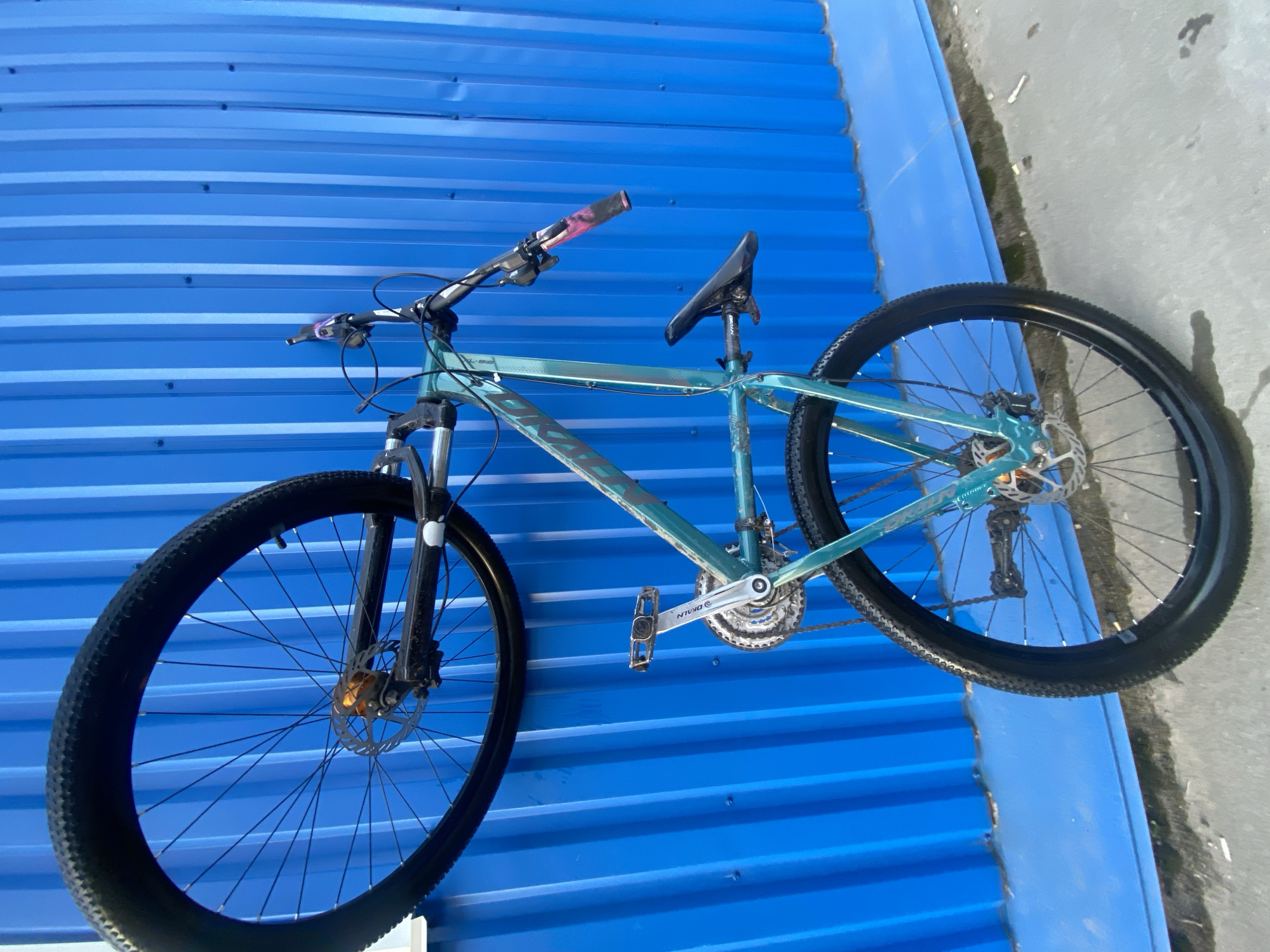 A teal-colored mountain bike is resting against a blue corrugated metal wall. The bike is oriented diagonally across the frame, with the front wheel on the left and the rear wheel on the right. The bike has black tires, a black seat, and a drivetrain consisting of a chain, gears, and pedals. The handlebars have pink grips. The location is Chaikovsky, Russia. Adjacent to the blue wall is a gray concrete surface with a patch of moss.