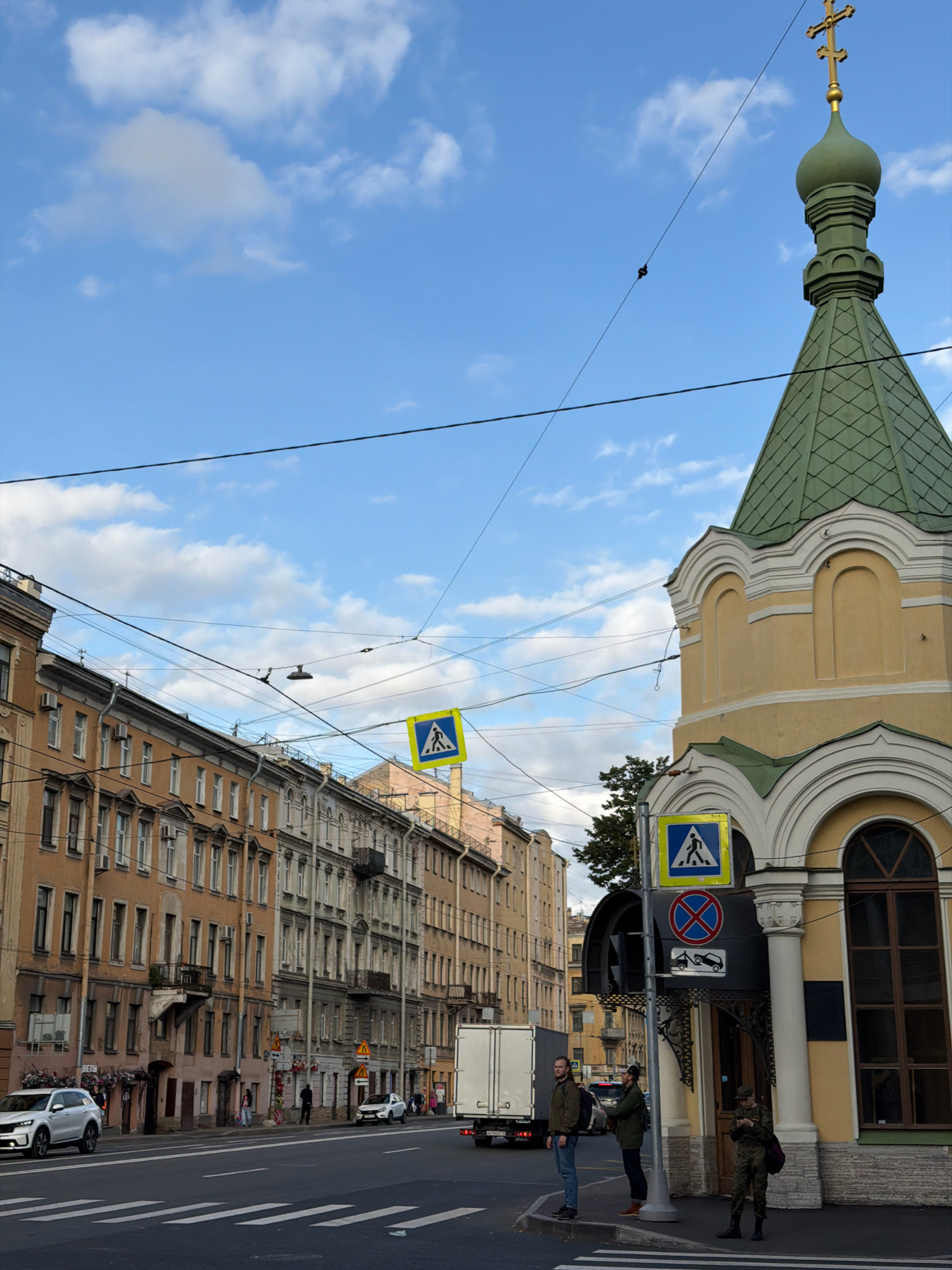 The image shows a street scene in Saint Petersburg, Russia. A multi-story building with ornate architecture, painted in yellow and green, stands on the right side of the frame. A green domed cupola with a gold cross tops the building. Across the street, a row of tall, older apartment buildings with cream and orange facades lines the left side of the road. Overhead, a complex network of electrical wires crisscrosses the sky. Pedestrian crossing signs, one with a blue background and a white pedestrian symbol, and another with a red circle and cross over a car symbol, are visible. Three individuals are standing on the sidewalk near a traffic light pole. One person, wearing a green jacket and jeans, stands facing left, while another, in a dark coat and hat, faces right. A third person, in camouflage uniform, is looking down at their phone. A white SUV is parked on the left side of the street, and a white truck is in the right lane. Pedestrian crosswalk markings are visible on the asphalt road.