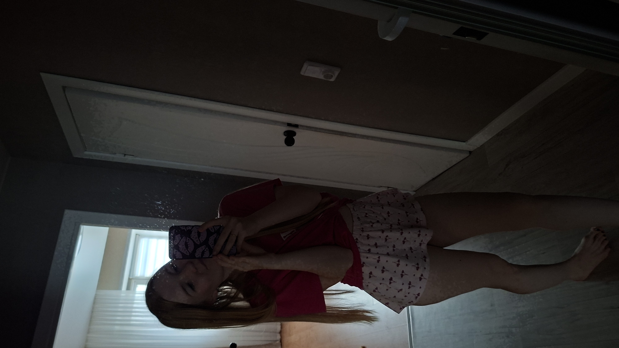 This is a vertical, full-body mirror selfie of a young woman. She is wearing a red t-shirt and a white skirt with a small red pattern. She has long blonde hair and is holding a phone with a purple case that has a lip pattern on it. The background appears to be a bathroom with a large mirror. There is a door with a white frame behind her, and a window with blinds is visible in the reflection. The lighting is dim, suggesting it might be evening or nighttime. The woman's expression is neutral.