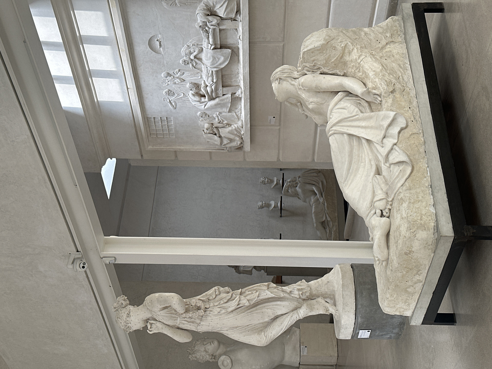 The image depicts a museum interior, likely an art gallery, with several classical-style sculptures. In the foreground, a white marble sculpture of a woman reclining on a stone chaise lounge is prominently displayed. She appears to be asleep or in a state of repose, with her head tilted back and her eyes closed. Her body is draped in flowing robes.
Behind this central figure, to the left, stands another tall white marble statue of a woman in a dynamic pose, her hand raised to her face. To the right of the reclining figure, a large relief sculpture adorns the wall, depicting a scene with multiple figures surrounding a central figure lying down, possibly a depiction of death or a significant event.
In the background, other sculptures are visible, including busts on pedestals and another reclining figure in a more distant alcove. The environment appears to be well-lit, with natural light filtering in from a skylight or a series of windows in the ceiling, casting geometric patterns of light and shadow on the floor and walls. The walls themselves are a neutral, light color. A security camera is visible mounted on a track near the ceiling. The floor is a polished, light-colored stone or concrete. There are no people actively interacting with the sculptures, suggesting the image was taken during a time when the gallery was closed or sparsely populated.
There is no information in the image to suggest the location is in the city of Caniezza, Italy.