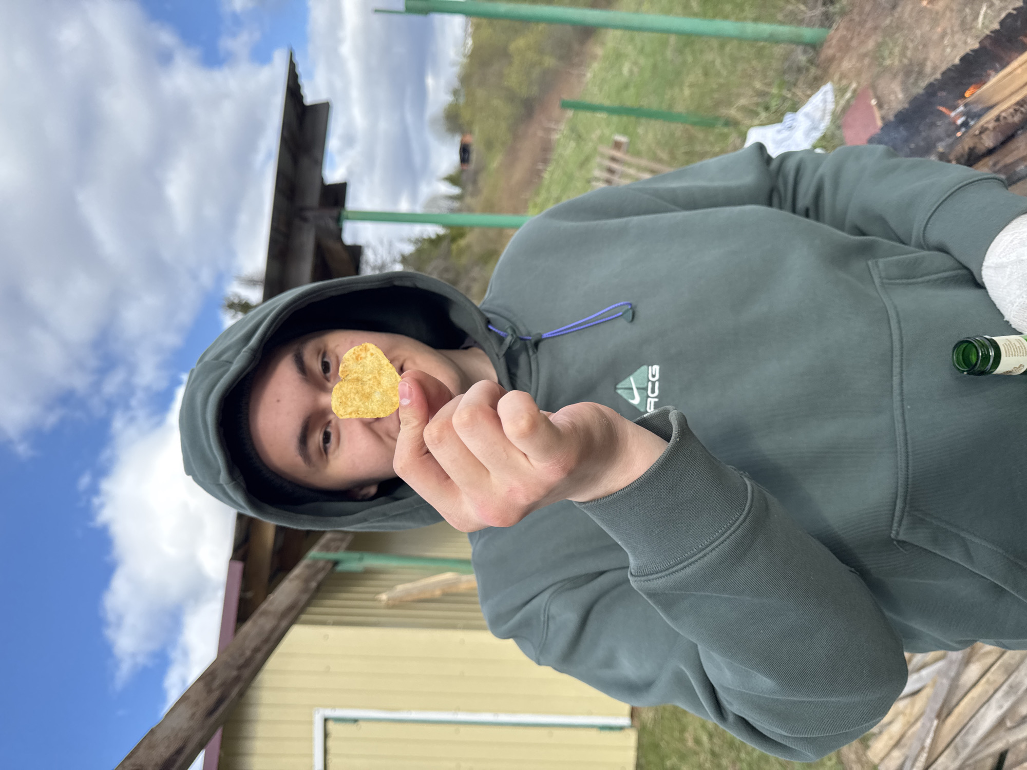 A person wearing a dark green hooded sweatshirt is holding a potato chip shaped like a heart in front of their face. The location is outdoors, with a small yellow building visible in the background. There's a patch of grass and what appears to be a fence or railing. A fire is burning nearby. The sky is partly cloudy. The location is not confirmed to be Abrosy, Russia.