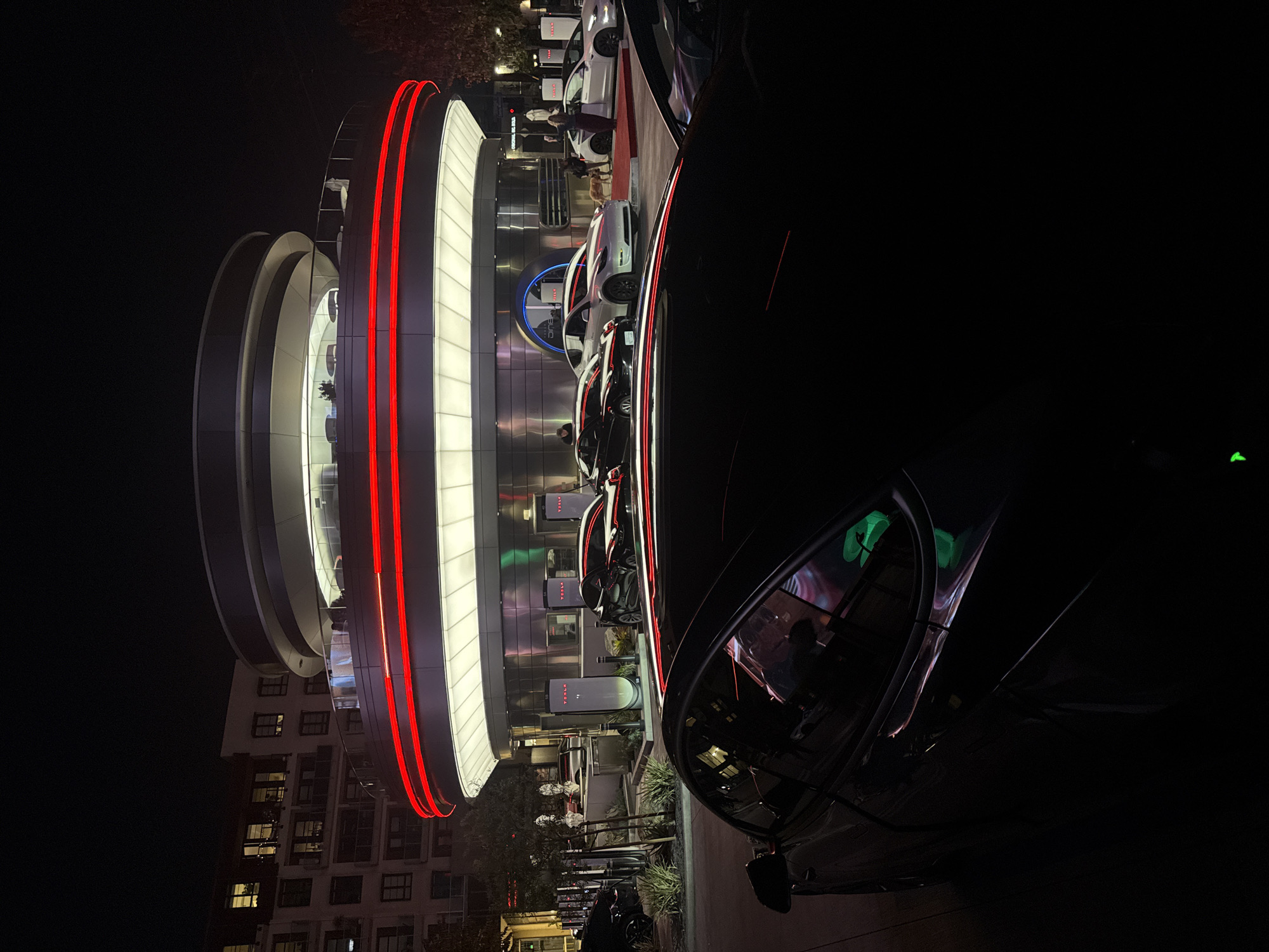 The image shows a Tesla charging station in West Hollywood, United States, at night. The station has a futuristic design with illuminated, concentric rings that dominate the left side of the frame. Red neon lights outline some of the rings. Several Tesla cars are plugged into the charging stations in front of the main structure.
On the right side of the frame, a dark-colored vehicle, possibly a car, is prominently positioned. The building is in a mostly urban setting, with residential buildings visible in the background.
