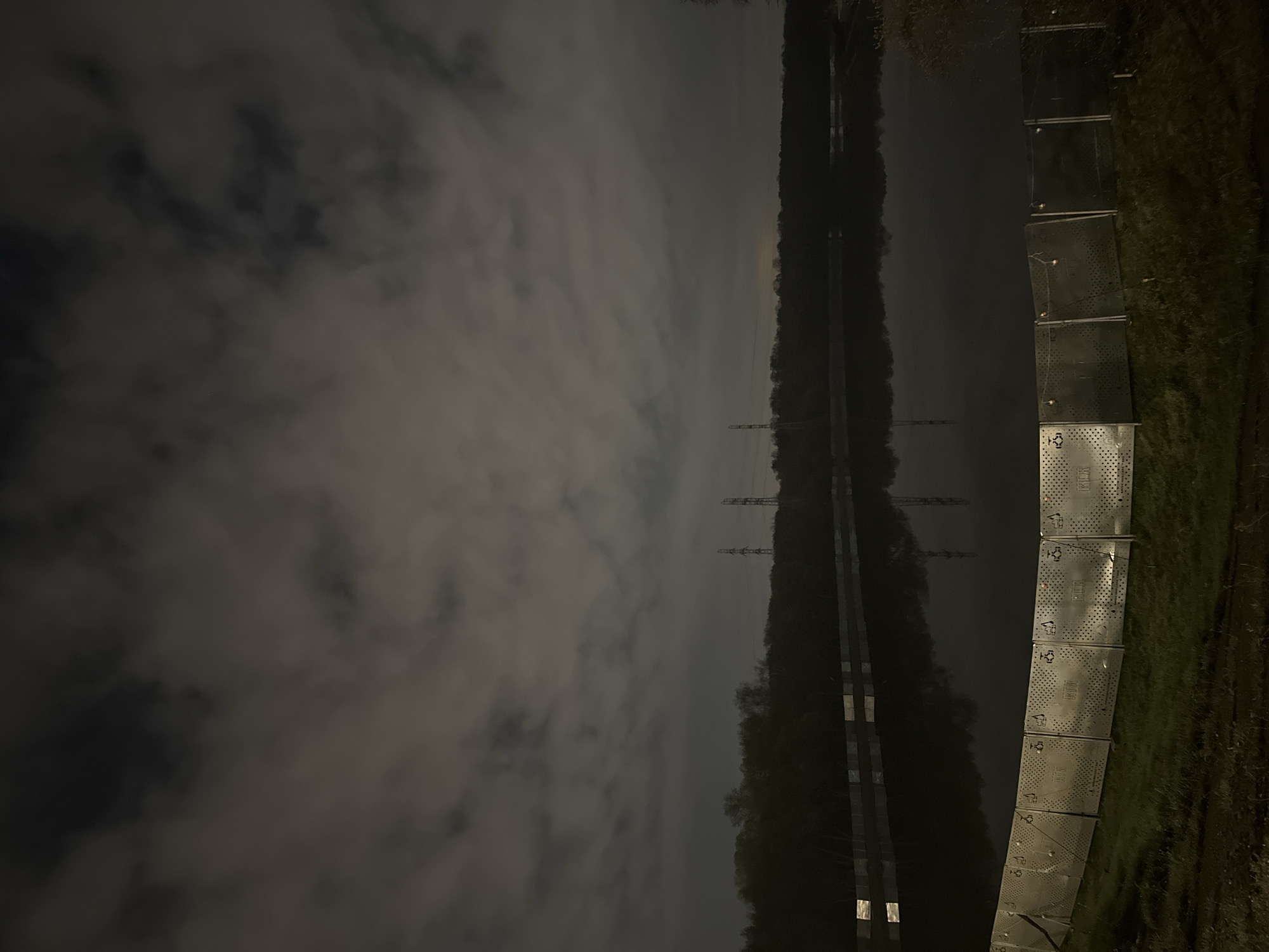 The image depicts a landscape at night. A canal or river runs through the center of the image, bordered by trees and foliage. Power lines are visible in the distance, crossing over the water. To the right of the frame, a series of metal barriers or containers are lined up on a grassy area. The sky is overcast, with thick clouds obscuring the view. The photo appears to be taken at an angle, with the ground sloping upwards from the lower left to the upper right. The exact location in Moscow, Russia, cannot be determined from this image alone.