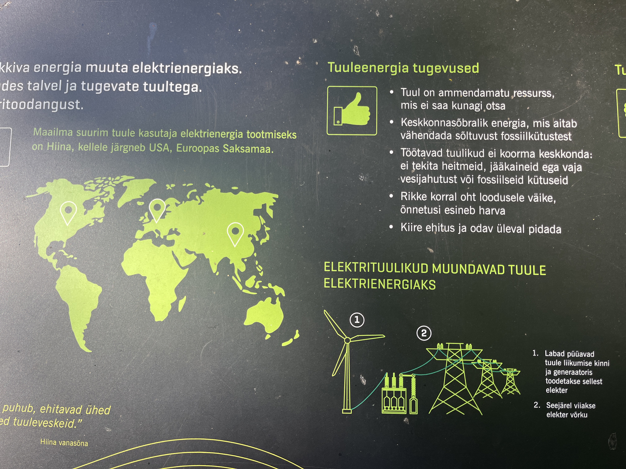 This image is a photograph of a display panel or information board, likely in an outdoor setting given the naturalistic background and potential for public viewing. The primary subject is an educational exhibit about wind energy. The display features a world map in green, highlighting China, the USA, and Germany as major wind energy users.
To the right of the map, there is text in Estonian titled "Tuuleenergia tugevused" (Strengths of Wind Energy), listing several bullet points about the benefits of wind power, such as it being an inexhaustible resource, environmentally friendly, and quick to build. A green thumbs-up icon is also present next to this section, reinforcing the positive aspects.
Further to the right, there's a section with the heading "ELEKTRITUULIKUD MUUNDAVAD TUULE ELEKTRIENERGIAKS" (Wind Turbines Convert Wind to Electricity). This section includes a simplified diagram illustrating the process: a wind turbine (labeled with '1') connected to a transformer and then to power lines (labeled with '2'). Accompanying text explains how the wind turbine's blades capture wind motion, a generator produces electricity, and this electricity is then fed into the grid.
At the bottom left, there's a fragment of text that appears to be a proverb or saying: "puhub, ehitavad ühed...ed tuuleveskeid." (blows, some build... windmills). This is attributed to "Hiina vanasõna" (Chinese proverb).
The overall tone of the display is informative and promotional, aiming to educate viewers about the advantages and mechanics of wind energy. The lighting suggests it's daytime, and no specific weather conditions are evident beyond the subject matter of wind. The location context provided (Toila, Estonia) implies this is likely a public space, possibly a park, educational center, or near a wind farm, designed for public engagement with renewable energy topics.