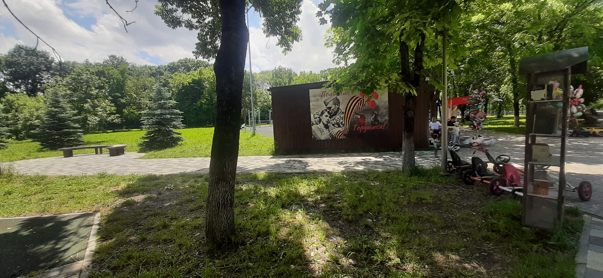 A tree stands in the foreground of this outdoor scene. To the left of the tree, a paved area with a bench is visible. Behind the bench, there are coniferous trees and lush green grass. To the right of the tree, a building with a large mural is present. The mural depicts soldiers and is adorned with a ribbon and red flowers. Further to the right, several people are seated on benches, and a collection of balloons is displayed. In the distance, more trees and a park-like setting can be observed. There are also several pedal cars parked in the area. The sky is partly cloudy. The overall setting appears to be a public park in the city of Yessentukskaya, Russia, likely decorated for a commemorative event given the mural's imagery.
