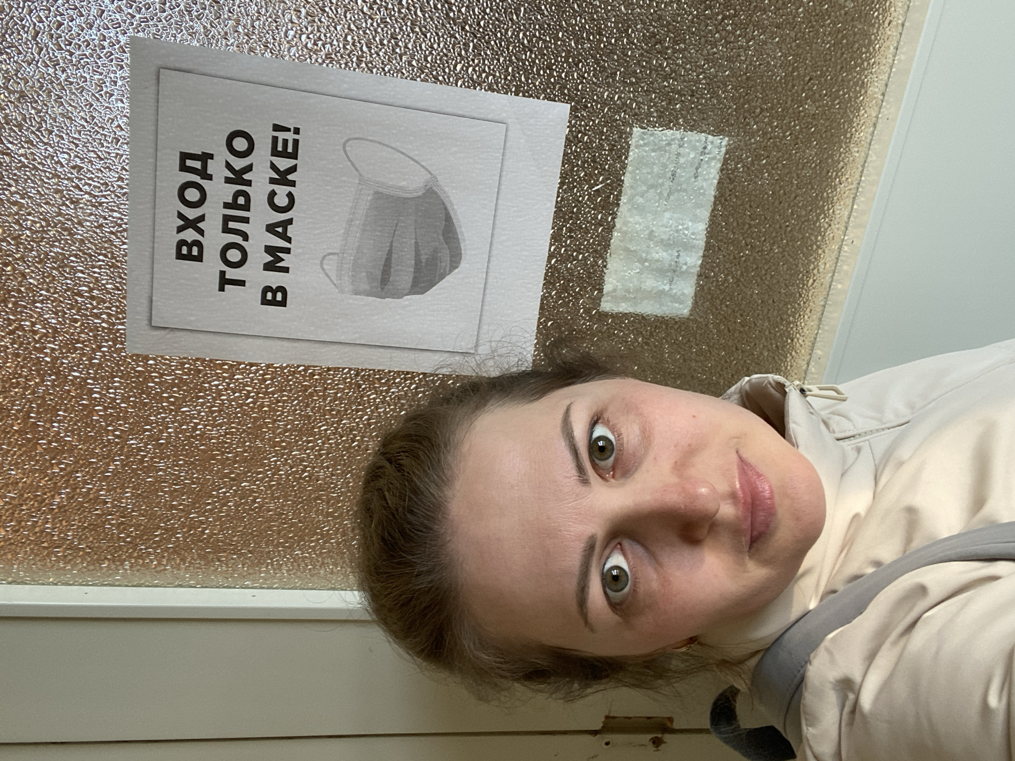 A woman stands in front of a door with frosted glass and a white frame. She wears a light beige jacket with a grey shoulder strap visible. A sign is taped to the glass of the door with the text "ВХОД ТОЛЬКО В МАСКЕ!" (Entrance Only with Mask!) and a picture of a face mask. The image was likely taken indoors. The specific location is Марковский, Russia.