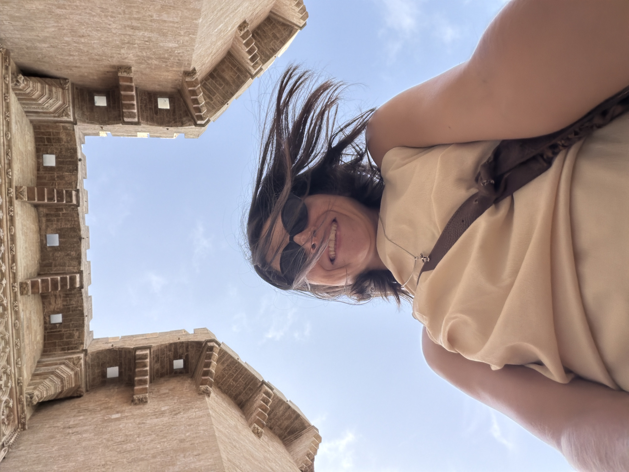 A low-angle, wide shot captures a woman in the foreground, looking up at the camera with a wide smile. She has dark hair that is windblown and she is wearing sunglasses and a light-colored, sleeveless top. A brown strap crosses her chest, and a delicate silver necklace with a cross pendant is visible. Behind her, the clear blue sky is framed by ancient stone architecture. The architecture appears to be a large, stone structure with intricate detailing and squared openings, possibly a historic building or tower. The perspective suggests the photo was taken from ground level, looking upwards into an archway or between two towering structures.