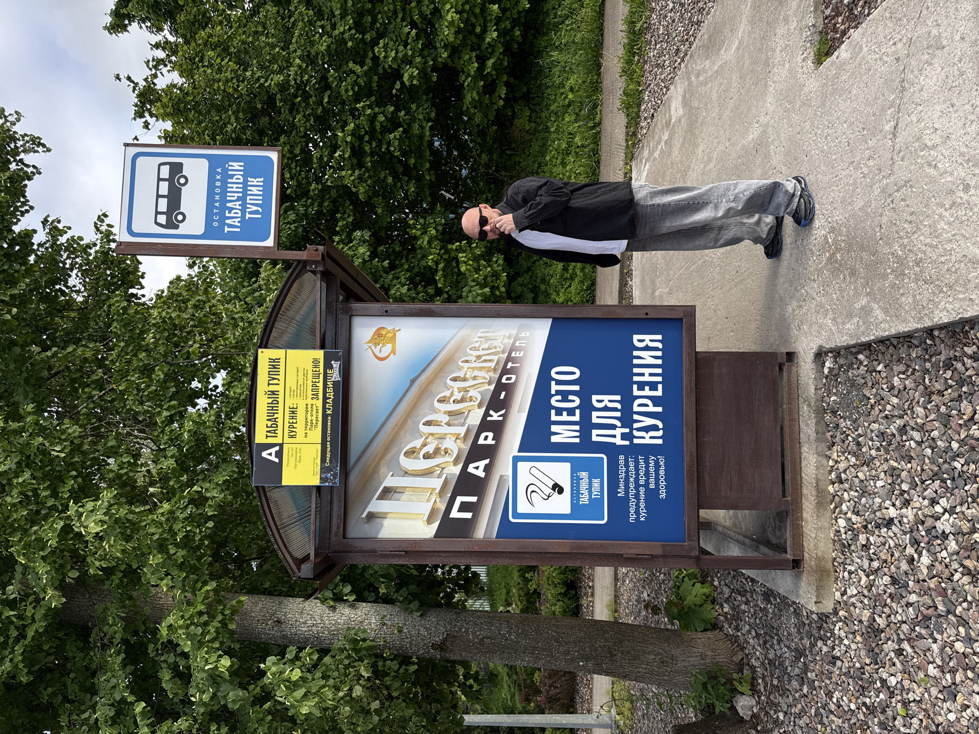 A man in sunglasses, a black jacket, and grey pants stands at a designated smoking area in Peresvet, Russia. The location is marked with signs in Russian that translate to "Tobacco Dead End," including one resembling a bus stop sign. The area is adjacent to a hotel called "Park-Hotel Peresvet." A sign also warns about the health risks of smoking. The man appears to be using his cell phone. The environment includes trees, a concrete walkway, and loose stones on the ground.