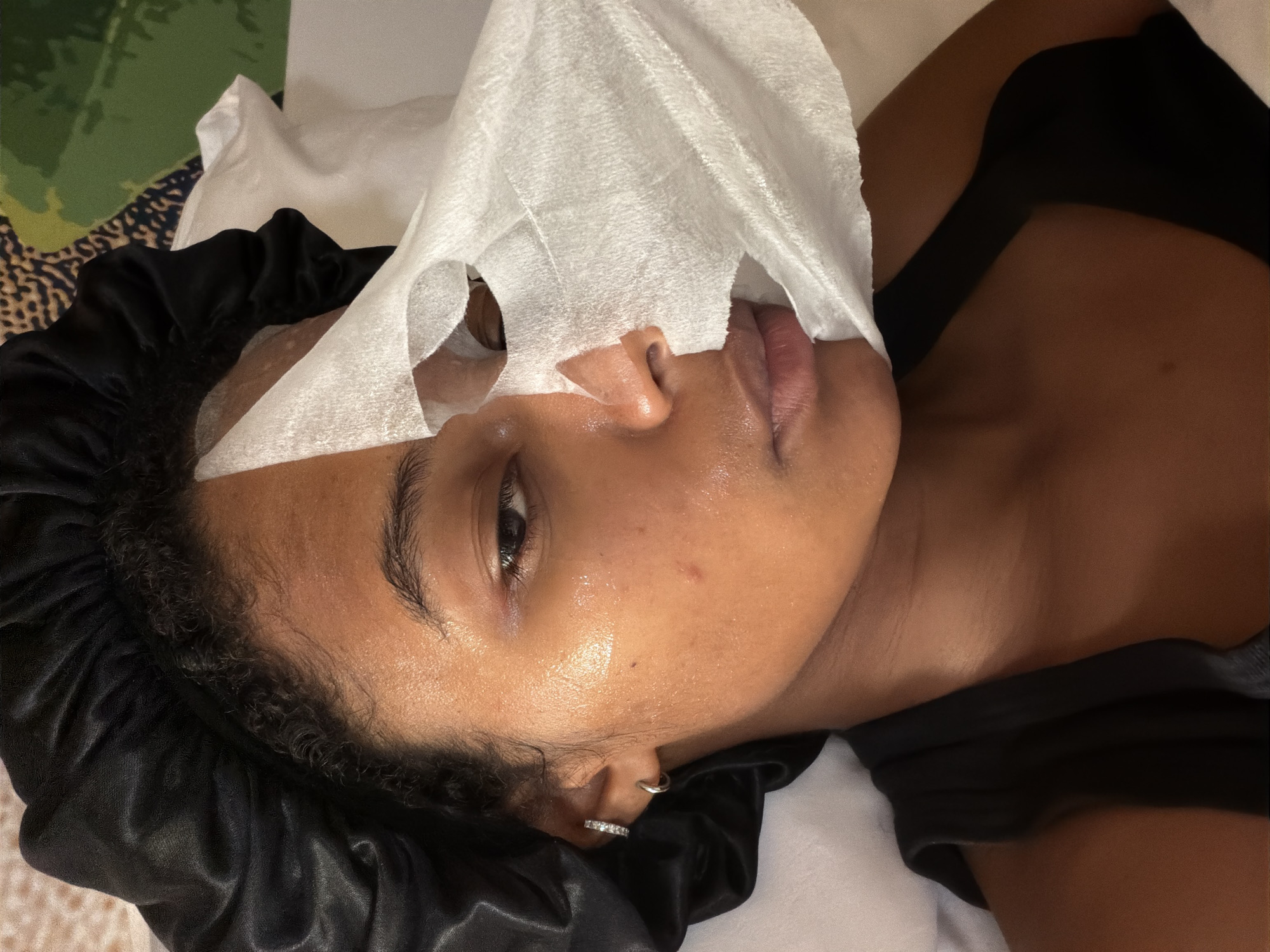 A close-up, over-the-shoulder shot shows a person lying down, wearing a black satin bonnet and a black tank top. A white sheet mask is partially applied to their face, covering the right side of their face and extending towards their hair and shoulder. The person's left eye is visible, with a subtle sheen on their skin. They are wearing small hoop earrings and a studded earring in their left ear. The background shows a textured surface, possibly a bedspread or wall covering, with green and beige abstract patterns. The image is a selfie-style portrait, likely taken indoors. There is no discernible location in Bremen, Germany.
