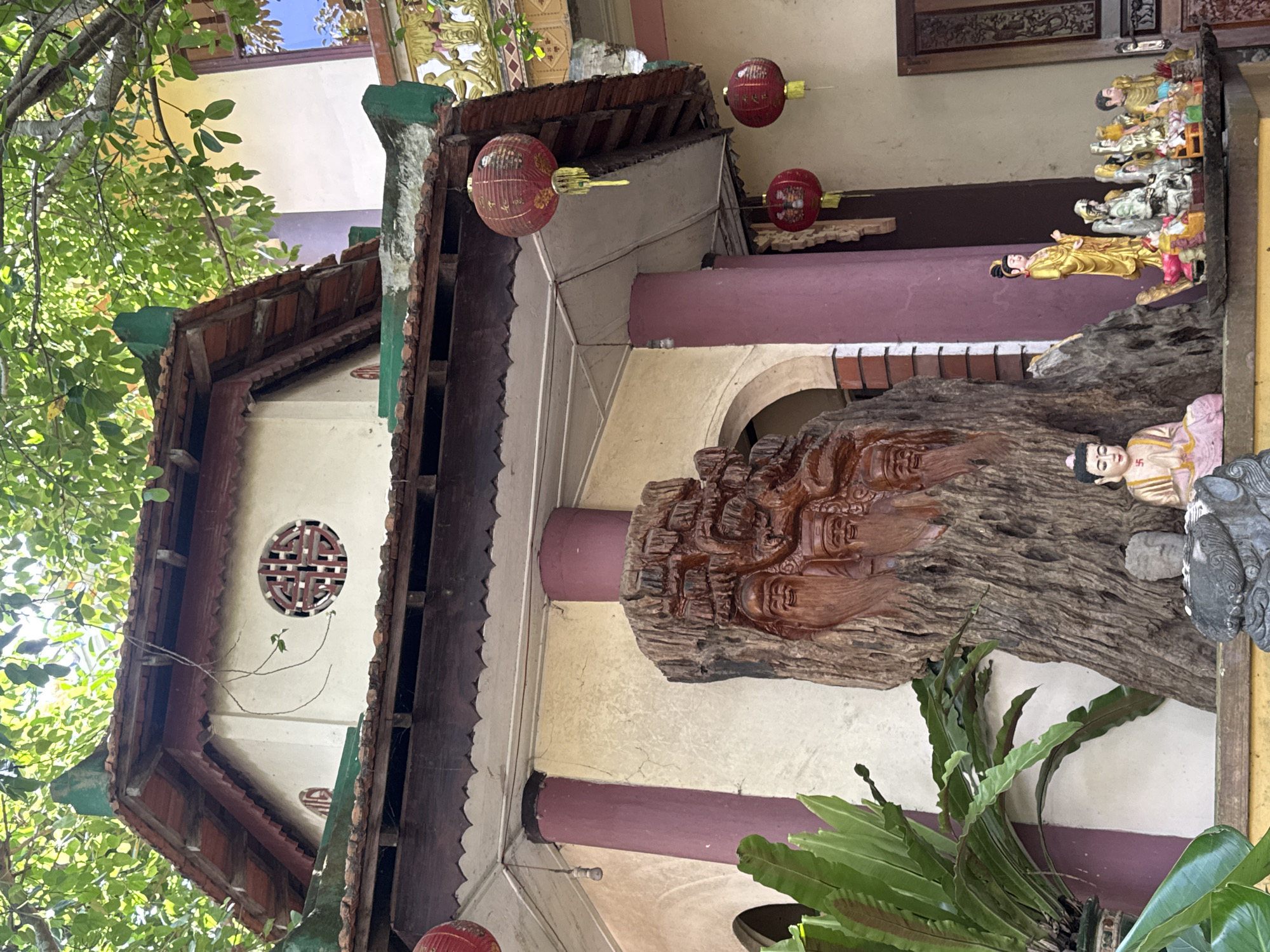 The image depicts the exterior of a building with distinctive Vietnamese architecture, likely a temple or shrine, in Da Lat, Vietnam. The structure features a tiered, tiled roof with green accents and intricately carved wooden eaves. A circular window with a geometric pattern is visible on the cream-colored wall. Red, spherical lanterns with yellow tassels hang from the eaves, adding a festive touch.
In the foreground, a large, intricately carved wooden trunk stands as a central focal point, featuring carved faces with beards and expressions of serenity. To the right of the trunk, a collection of small statues, including a golden figure of a woman in traditional attire and other deities, are displayed on a shelf. A smaller seated Buddha statue with hands clasped in prayer is positioned in front of the wooden trunk. Green foliage from a large plant is visible to the left, and part of another plant is to the right of the trunk. The overall scene is bathed in sunlight filtering through overhead tree branches.