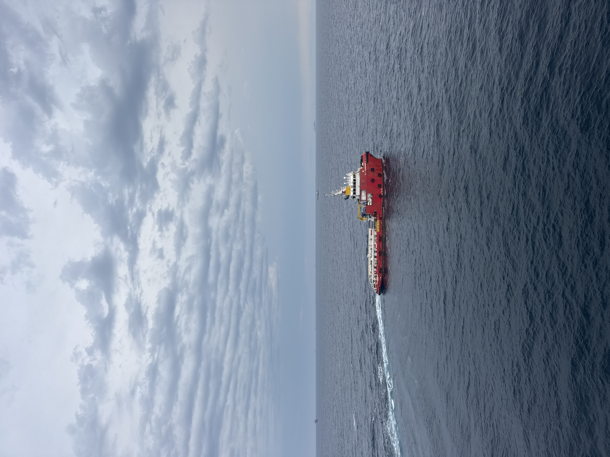 A bright red and white vessel, identified by the text "SEA CHAMPAGNE" on its side, navigates through a dark blue ocean under a cloudy sky. The ship is positioned in the lower right quadrant of the image, leaving a white wake behind it. Several other distant vessels and structures, possibly oil rigs, are visible on the horizon. The sky is filled with dense, grey clouds, creating an overcast and somewhat dramatic atmosphere. The water is choppy with small waves.
