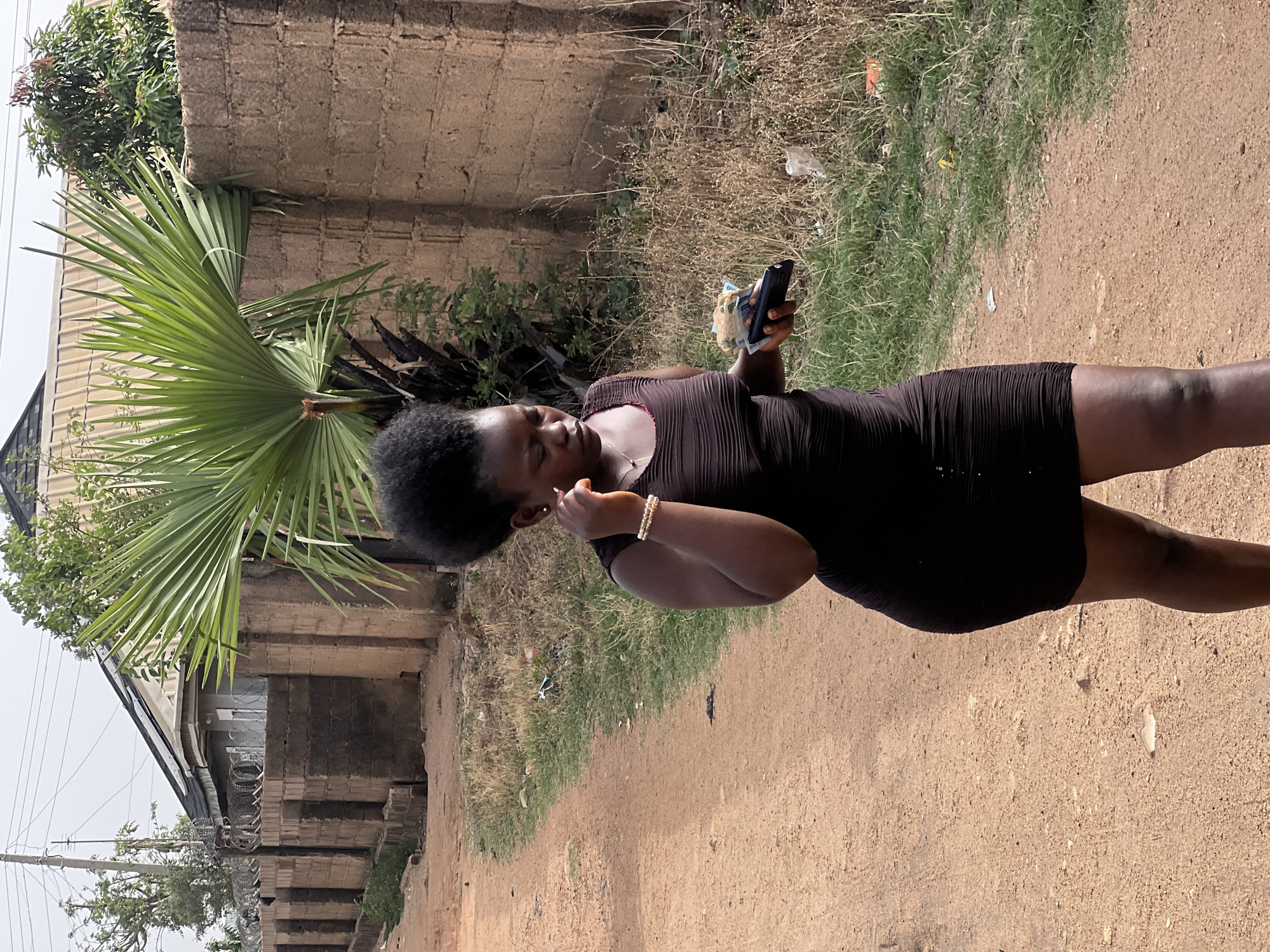 A medium-shot depicts a dark-skinned woman standing on a dirt path outdoors in Bukuru, Nigeria. She is wearing a short, dark-colored dress. She is facing towards the left, holding a phone in her left hand and touching her face with her right hand. She is wearing a bracelet on her right wrist.
The surrounding environment shows a dirt path with some sparse vegetation on either side. There is a brick or concrete wall and a palm tree to the left, along with a building with barbed wire on top of the wall. The sky is overcast.