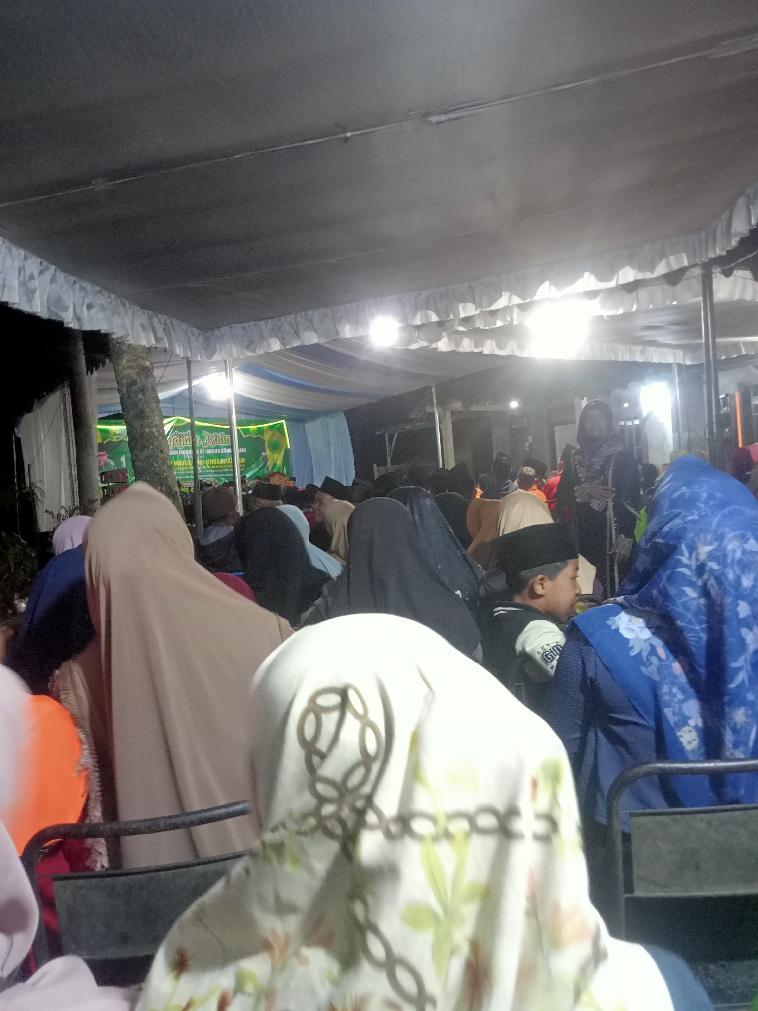 The image depicts an outdoor gathering under a large tent at night. The tent has a white fabric roof with decorative white ruffles along the edges. Several people are visible, most wearing Islamic attire, including headscarves and caps. In the background, a banner with Indonesian text and green and yellow accents is displayed, indicating an event. Lights illuminate the scene, creating a bright atmosphere. The foreground shows the backs of people seated or standing, with some wearing patterned hijabs. The overall impression is of a community event taking place in a populated area. The location is identified as Tanggaran, Indonesia.