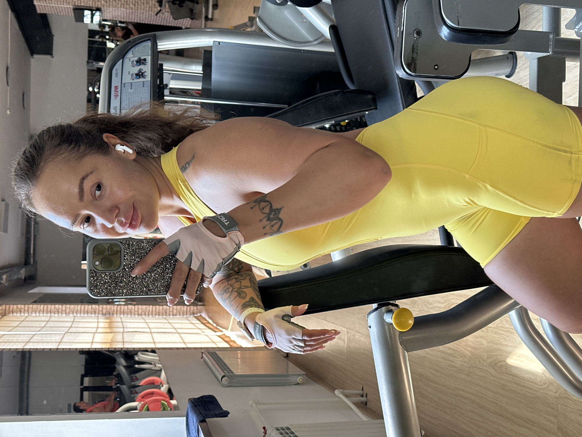 A woman is taking a mirror selfie in a gym. She is wearing a yellow workout outfit and light pink workout gloves. She has several tattoos on her arms. She is holding a phone with a glittery case. In the background, there is gym equipment and a reflection in the mirror shows more gym equipment and another person. The lighting suggests it could be daytime. The woman appears to be focused on taking the selfie.
