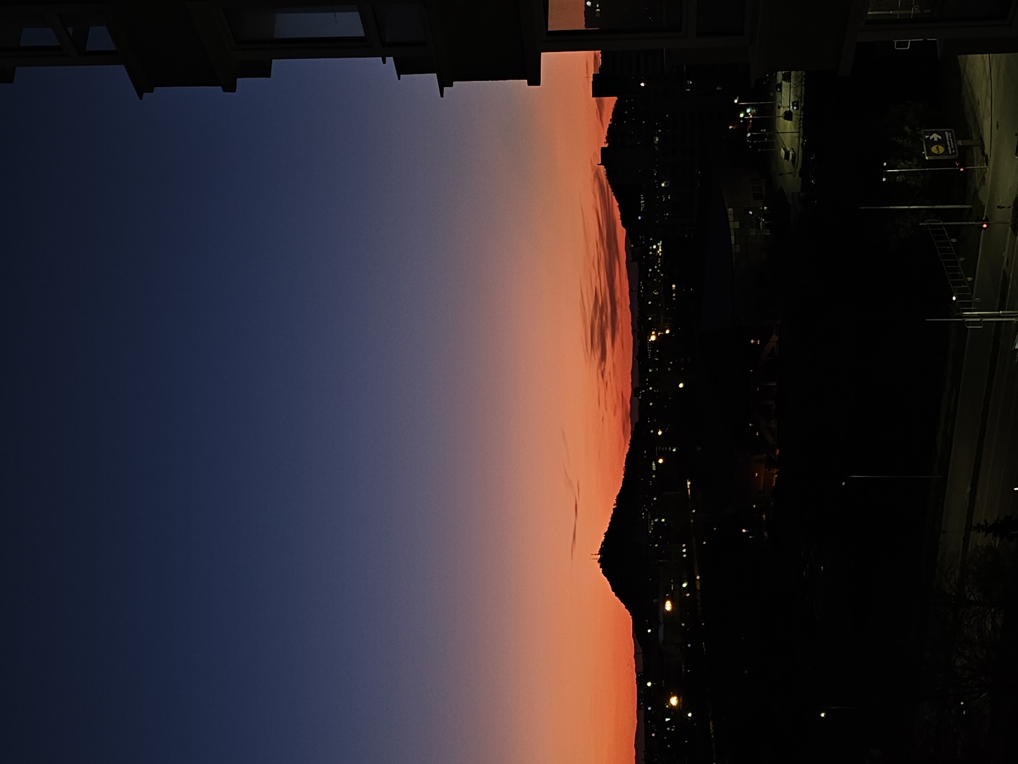The image depicts a view from an elevated position, possibly a building, at dusk or dawn, in Plovdiv, Bulgaria. The sky transitions from dark blue to a fiery orange near the horizon. A silhouette of a hill or mountain range is visible against the colorful sky. Below the horizon, there is a dimly lit urban landscape with scattered points of light, likely from streetlights and buildings. A road or street runs along the right side of the image, with a few visible street signs. A ladder leans against the side of the street. The upper portion of the image includes sections of what appears to be a building or architectural structure. No people are visible in the image.