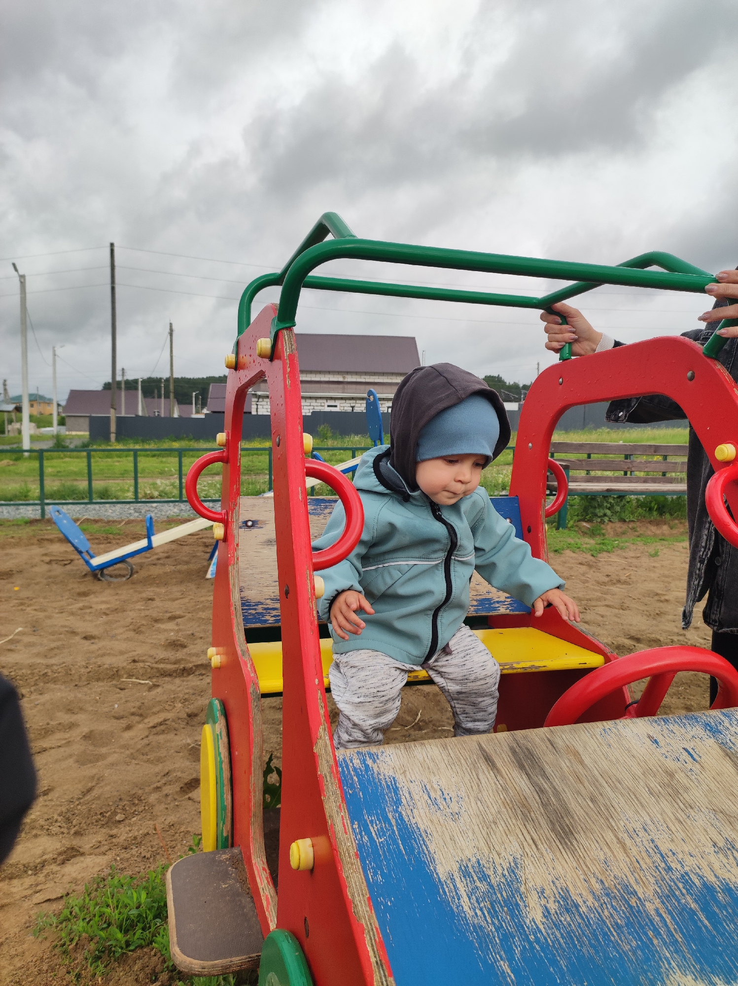 The image shows a young child playing on a playground in Фоки, Russia, under an overcast sky. The primary subject is a toddler, dressed in a teal hooded jacket with a dark hood and a light blue beanie, actively stepping into a wooden play structure designed like a car. The structure is painted in worn red, yellow, and blue, showing chipped paint. The child appears engaged and focused on their play.
To the right of the child, an adult's hands are visible, holding onto the green metal bars above the play car, possibly offering support or supervision. The playground area features sandy ground. In the mid-ground to the left, a blue and white seesaw is partially visible.
The background reveals a residential setting with several houses featuring dark roofs, separated by a green fence and grassy areas. Utility poles and power lines are visible against the cloudy, gray sky. The weather appears to be overcast, suggesting a cool day, consistent with the child's layered clothing. No visible text is present in the image.