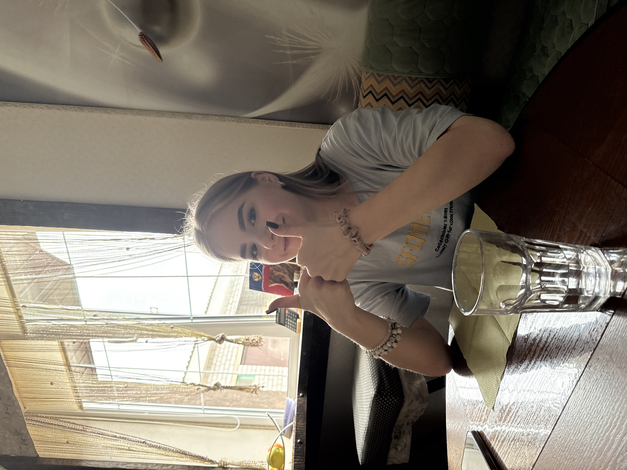 This indoor photograph, taken in Krapivka, Russia, features a young woman looking directly at the camera and giving two thumbs-up gestures, indicating approval or positivity. She has long, light-colored hair and is wearing a light grey t-shirt with partially visible text, including "SFODD," "Collectibles," "d Boxes," "th For," and "Good Frie." She accessorizes with a scrunchie-like bracelet on her left wrist and a white beaded bracelet on her right.
The woman is seated at a wooden table, where a clear glass rests on a light green placemat. Bright natural light floods the scene from a large window to her left, suggesting it is daytime. The window is adorned with intricate string or bead curtains, and a hint of the exterior is visible beyond. A small yellow object can be seen on the windowsill.
The background reveals an interior with varied wall coverings: a subtle textured pattern behind her head transitions to a more pronounced zigzag geometric pattern further down and to the right. A ceiling light fixture with a small decorative pendant is also visible at the top of the frame. The overall atmosphere is casual and cheerful.