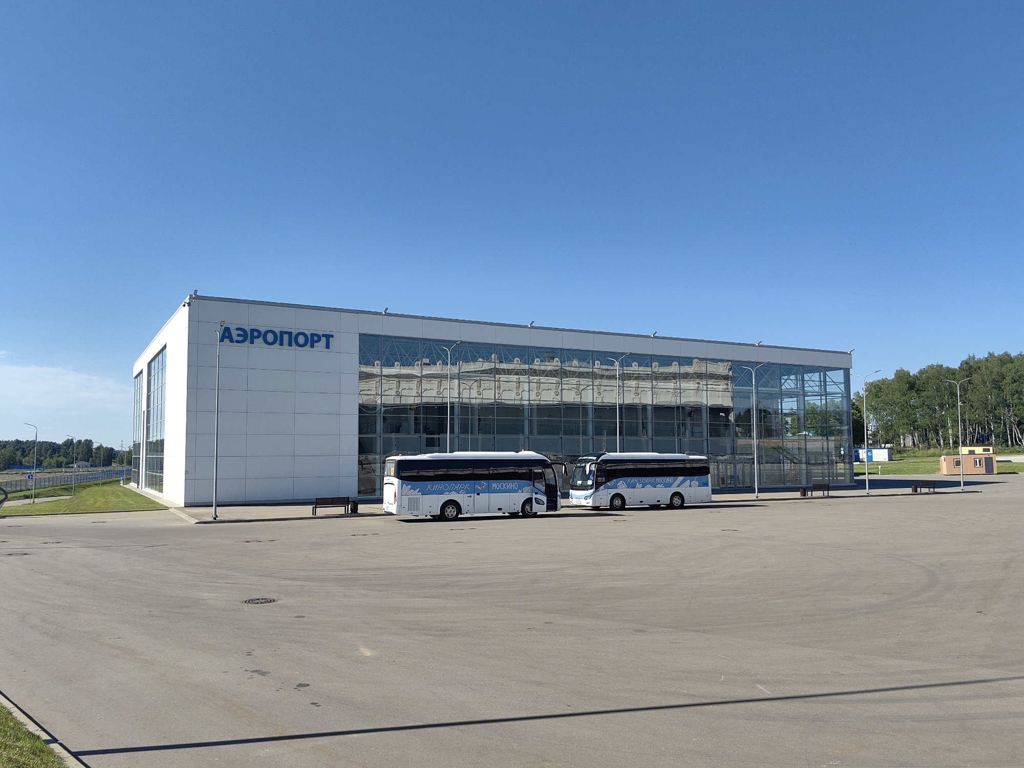 The image shows an airport terminal with the word "АЭРОПОРТ" (Airport) in blue letters on a white facade. The terminal has a large glass section reflecting the sky and trees. Two white buses with blue lettering are parked in front of the building. The buses have "КИНОПАРК" (Kinopark) and "МОСКИНО" (Moskino) written on them. The scene is set in a paved area with some benches and lampposts. In the background, there are trees and a clear blue sky.