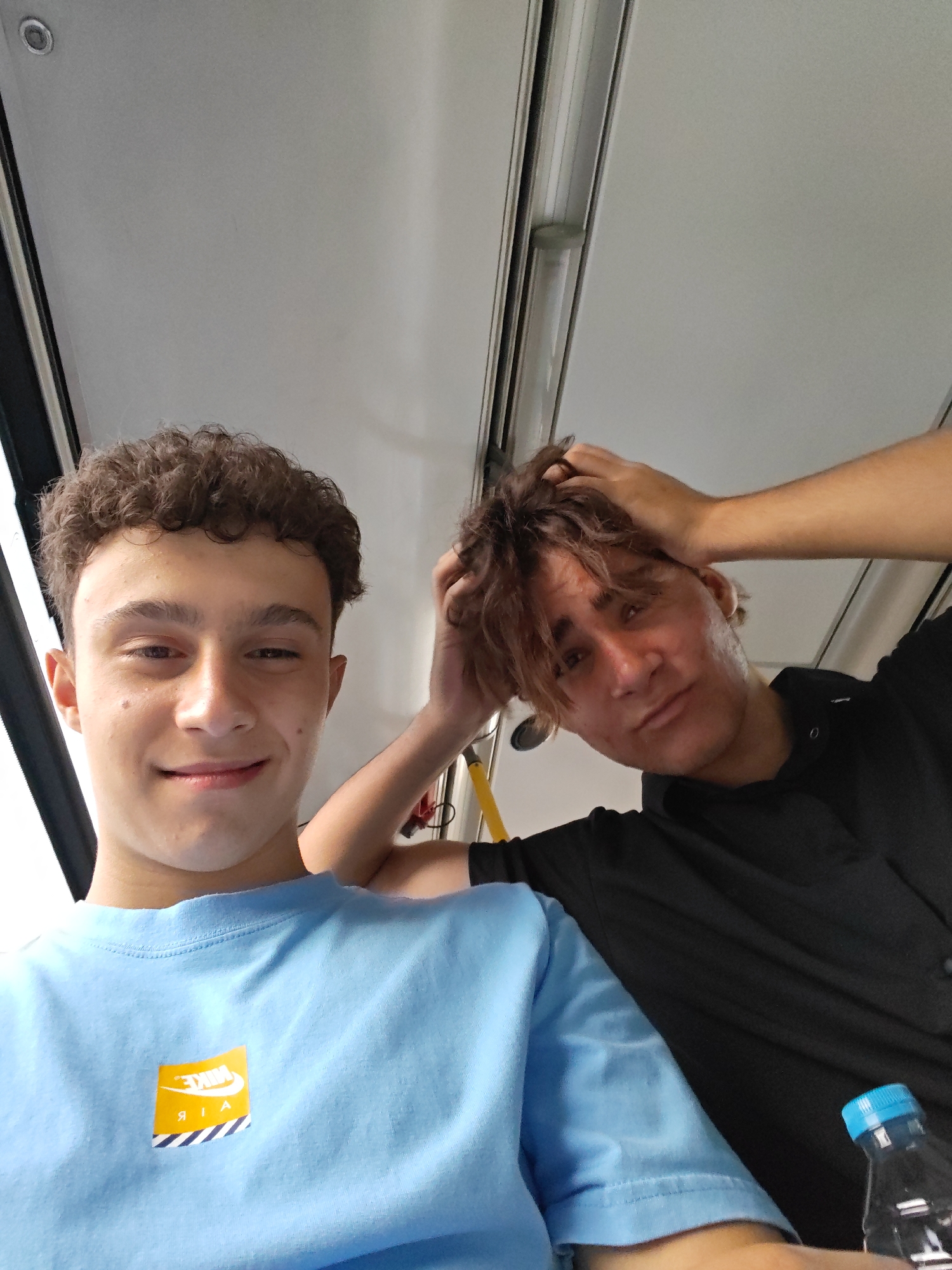 Two young males are taking a selfie indoors, possibly on public transport, given the overhead lighting and handrails visible. The male in the foreground, on the left, has curly brown hair and is wearing a light blue t-shirt with a yellow and white Nike Air logo. He is smiling and looking directly at the camera. The second male, on the right, has tousled brown hair and is wearing a black t-shirt. He is holding his hands up to his head, looking towards the camera with a relaxed expression. A clear plastic water bottle with a blue cap is visible on the right side of the frame. The lighting suggests an indoor environment, possibly during the day. There is no specific visual information in the image to indicate the location within the city of Stara Zagora, Bulgaria.