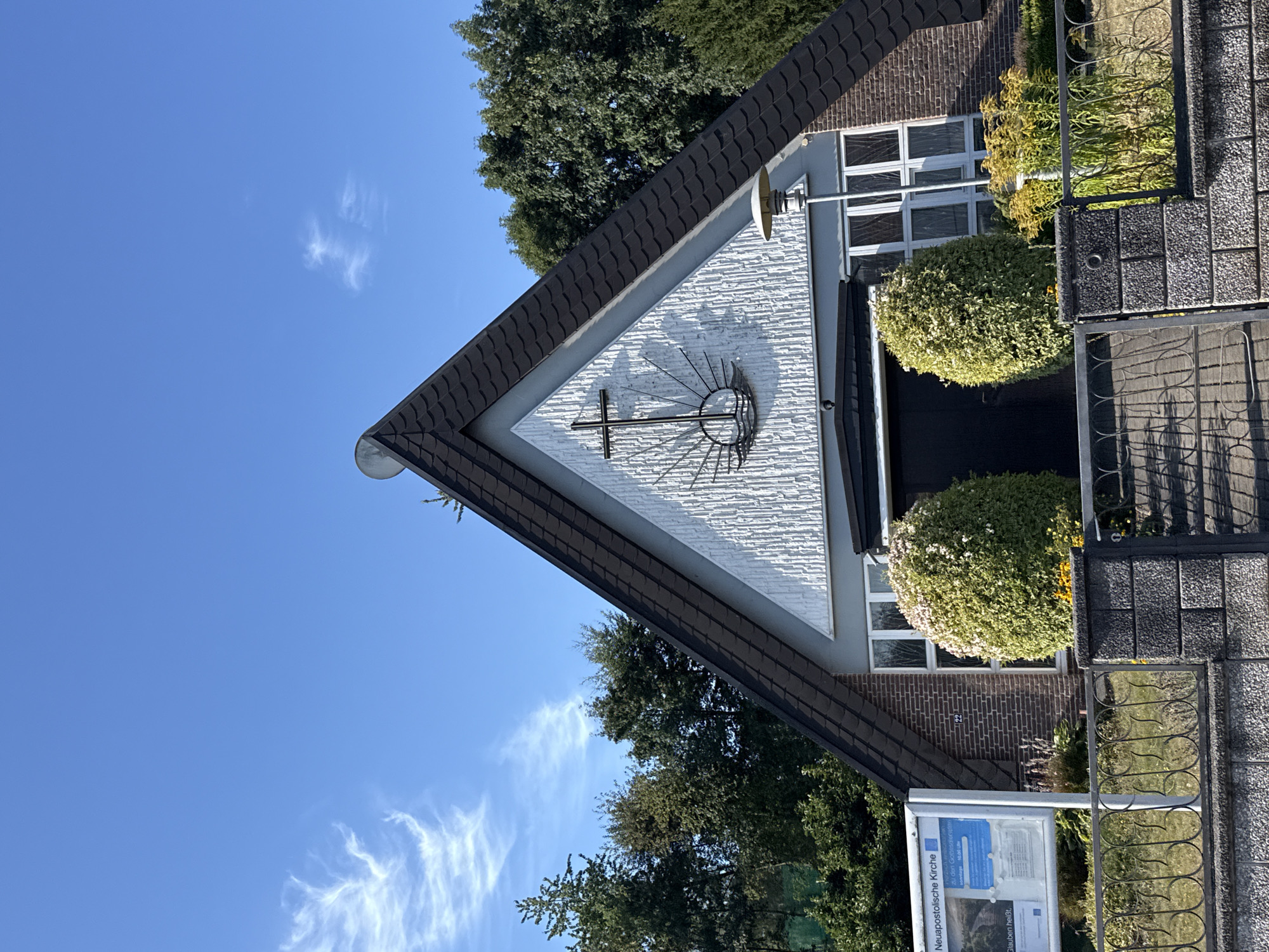 The image shows the exterior of a New Apostolic Church building in Bad Sachsa, Germany, on a clear, sunny day. The church has a distinctive A-frame roof with dark grey shingles. A white, textured facade forms the triangular gable, on which a dark metal cross with a radiating sun symbol is mounted. To the left of the entrance, a light blue sign with white text reads "Neuapostolische Kirche" and details service times. The building has several windows, including a large, multi-paned window above the entrance. Two spherical, well-maintained green bushes flank the entrance, with a flower bed of yellow and white flowers visible in front of the building. A black metal fence with an ornate design runs across the foreground, with a grey stone pillar and gate. Trees and foliage surround the church, indicating a location with greenery. The sky is bright blue with a few wispy white clouds.