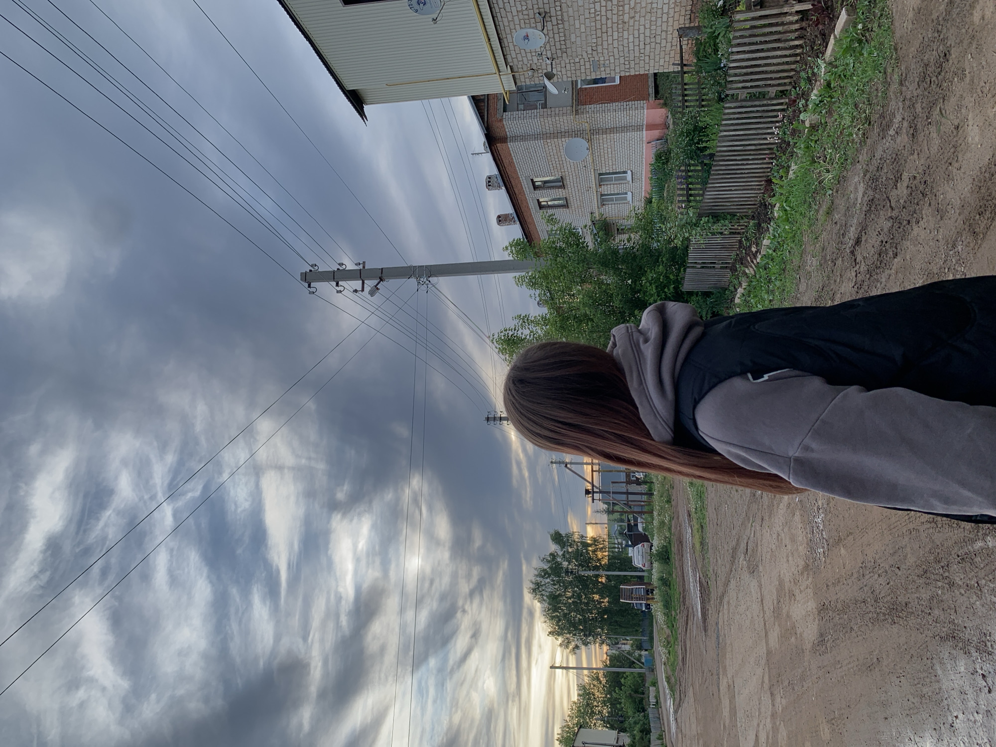 The image depicts a person looking towards a cityscape in Фоки, Russia. The view includes multi-story brick and corrugated metal buildings with satellite dishes, utility poles with power lines, trees, and a dirt road. The sky is filled with gray and white clouds.