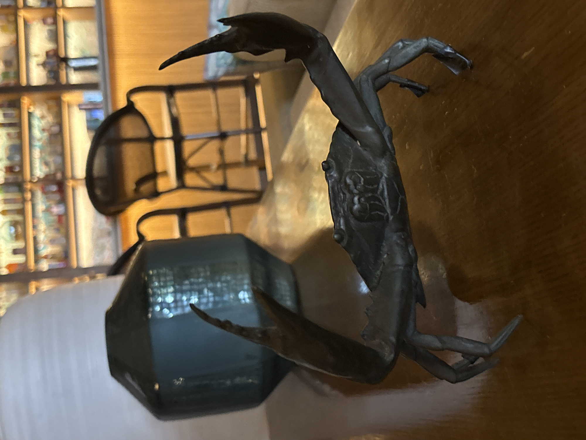 An ornate, dark metal crab figurine stands on a polished wooden surface. The crab's claws are raised, and its legs are positioned as if it is walking. Behind the crab, a large, teal vase with a rounded base and a tapered top sits on a light-colored textured surface. In the background, a bar area is visible with shelves stocked with bottles, illuminated by warm lighting. Two high stools with black seats and metal frames are placed in front of the bar. The scene appears to be indoors, possibly in a restaurant or lounge in Vagator, India, given the atmospheric lighting and bar setup.