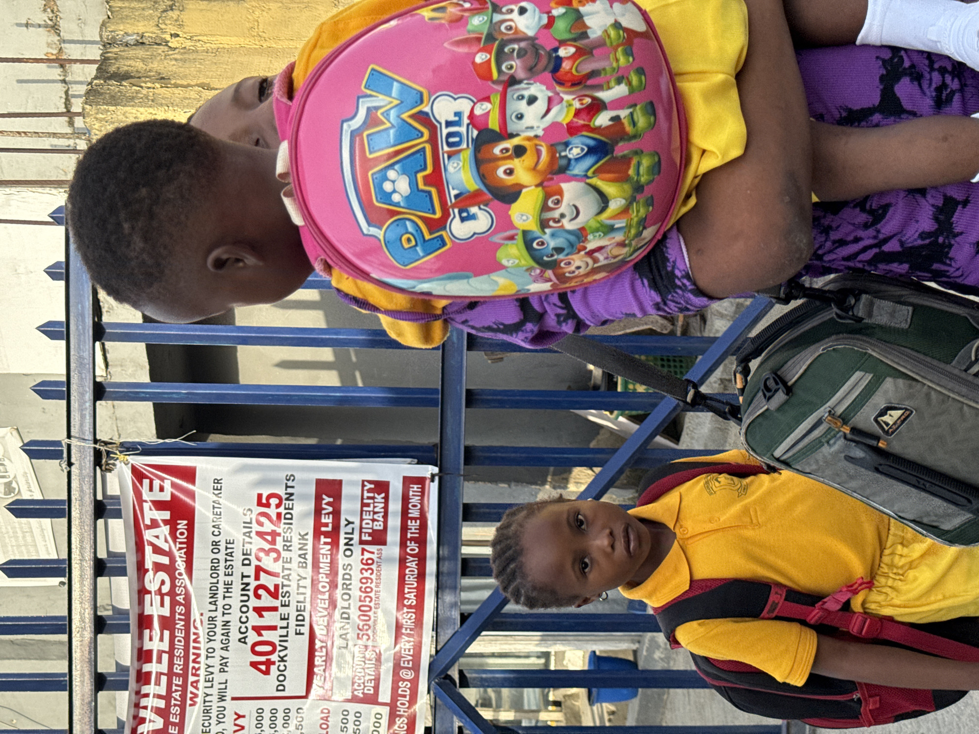 Two young children are visible in the image, seemingly near an entrance gate or fence. One child is being carried by the other. The child being carried has a pink "Paw Patrol" backpack. The other child is wearing a yellow shirt with a backpack. Attached to the gate is a sign for "Dockville Estate Residents Association", indicating the location is Dockville Estate. The sign lists payment details for the security levy and yearly development levy, with account details at Fidelity Bank. The sign also notes a meeting held every first Saturday of the month. The environment appears to be outdoors. The city is not specified as Ado.
