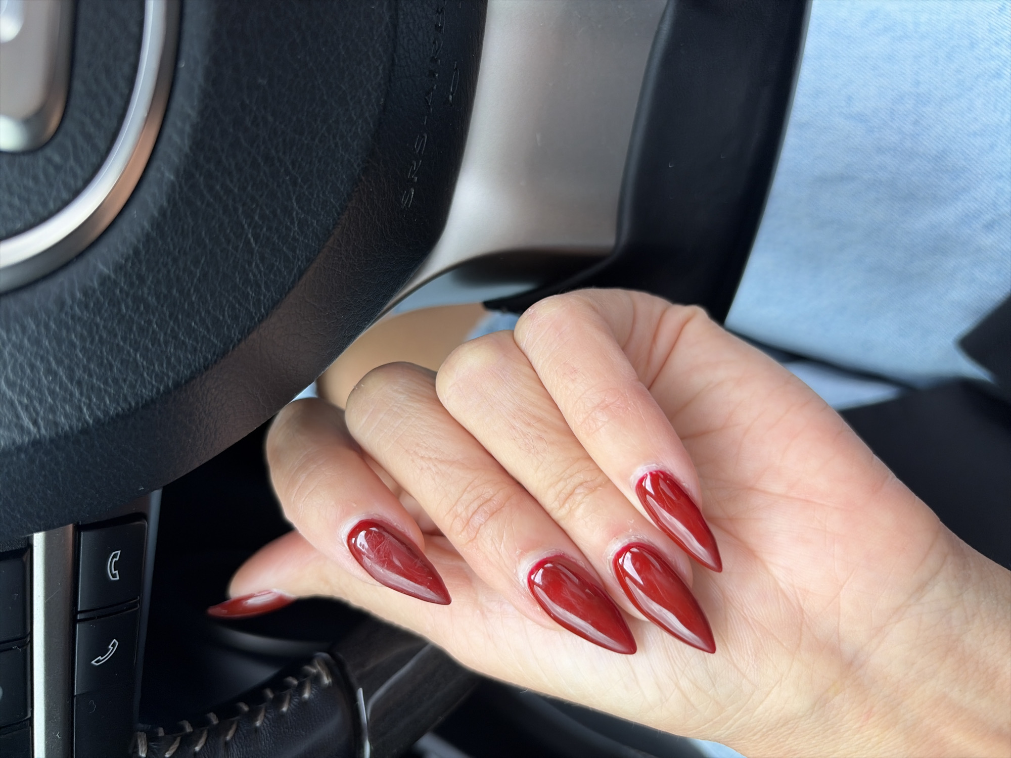 A close-up shot shows a person's hand with long, pointed, dark red acrylic nails. The hand is positioned near a black leather steering wheel, suggesting the person is inside a car. The steering wheel has visible stitching and a silver accent ring. Buttons with icons for making and receiving calls are visible on the steering wheel's control panel. The background is blurred, but a hint of light blue fabric, likely clothing, is present. The scene is indoors, within a vehicle, and there is no indication of the location being in Moscow, Russia.