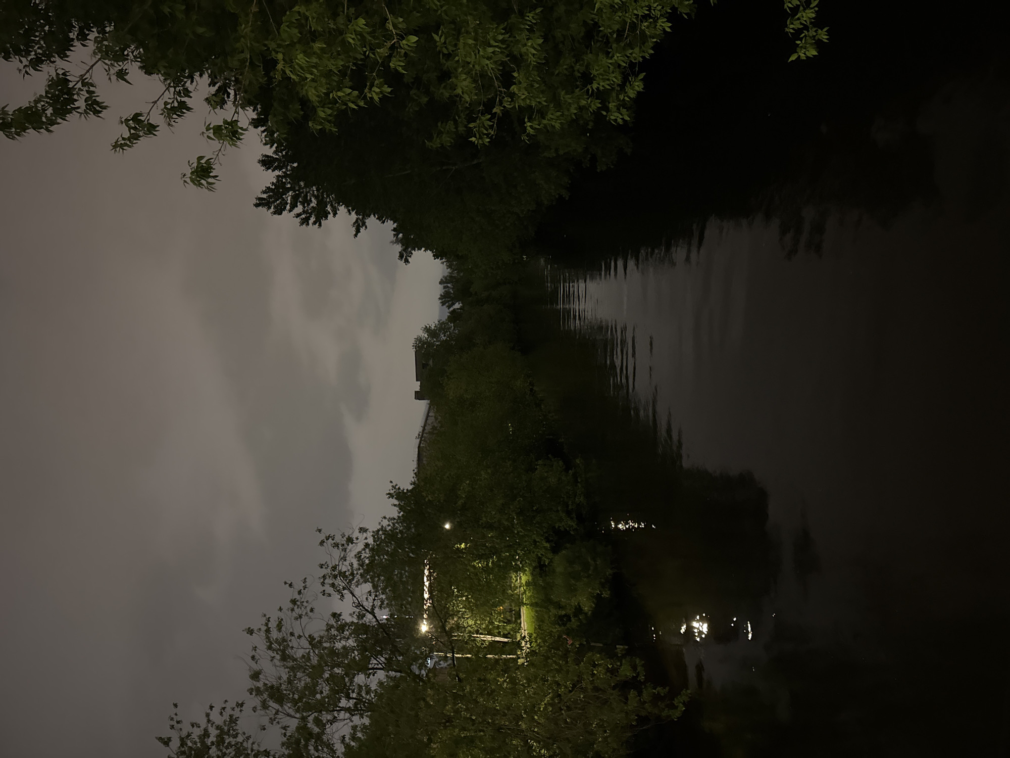 The image shows a dark canal in Saint Petersburg, Russia at night. The water reflects the sparse lighting along the canal. Trees line both sides of the canal, with their branches reaching overhead. The sky is overcast and cloudy. A building is visible in the middle distance. The overall scene is quiet and dimly lit.