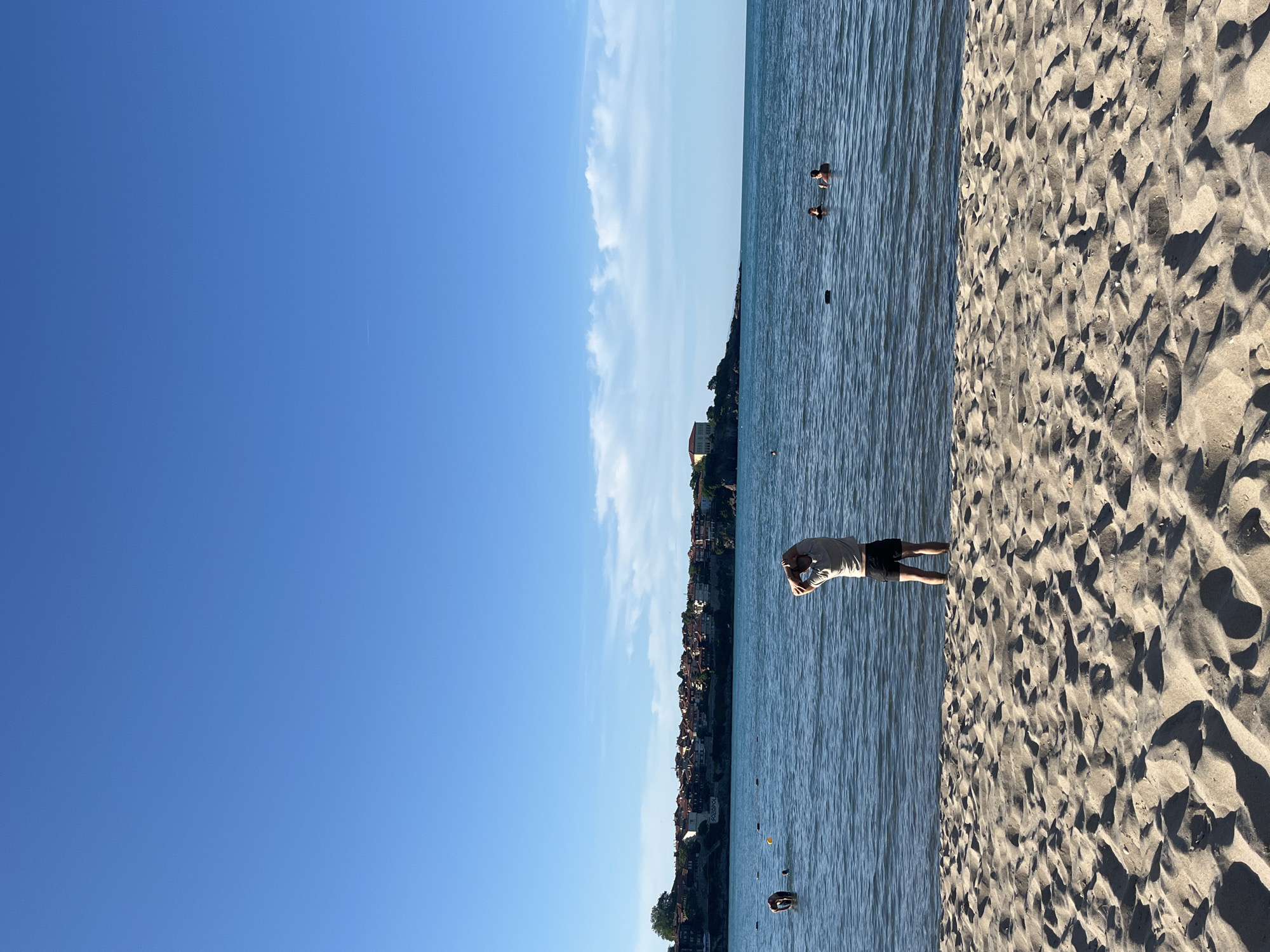 The image depicts a beach scene in Sozopol, Bulgaria, with the old town visible in the background. In the foreground, sandy beach stretches towards the sea, with ripples and shadows indicating a sunny day. The sea is calm and blue, with several people swimming. In the middle ground, a person stands in the water, facing away from the camera, with their arms raised behind their head, seemingly stretching. Further out in the water, other individuals are visible, some swimming and others wading. The background shows the picturesque old town of Sozopol, characterized by its red-tiled roofs and traditional architecture, situated on a hill overlooking the sea. The sky is a clear, bright blue with a few wispy clouds.
