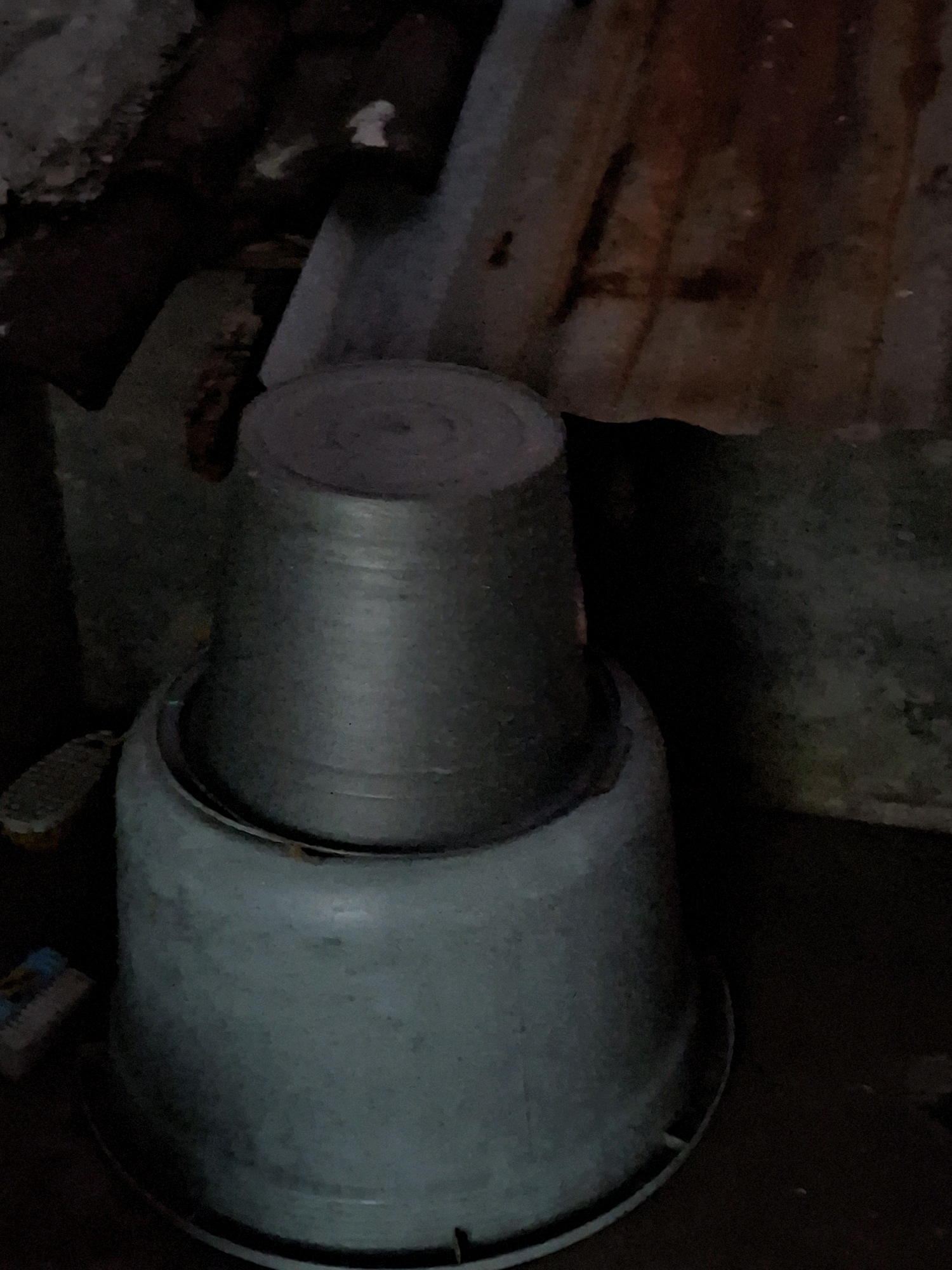 The image depicts two stacked metal pots, likely used for cooking or storage, situated outdoors. The larger, light blue pot is at the bottom, and a smaller, darker gray pot is placed on top. The pots appear weathered and are placed on a dark, uneven surface. In the background, there are elements of what appears to be a dilapidated structure, possibly a roof made of corrugated metal and wood, suggesting an outdoor storage or utility area. There are no people visible in the image. The scene appears to be in a rural or semi-urban setting in Lumajang, Indonesia, given the typical materials and the general ambiance of the environment, which often features open-air storage and utility spaces.