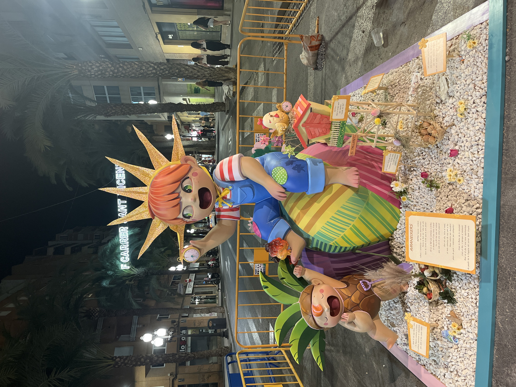 The image shows a nighttime street scene in Alicante, Spain, featuring a large, colorful artistic display. The display is a sculpture of a young boy with bright orange hair and a sunburst headdress, sitting on a large, striped sphere. He holds a small clock in his hand and is wearing overalls and a striped shirt. To his left, a smaller character, resembling a nut with a face, is reaching up. The display is surrounded by white gravel, and features miniature elements like a chicken coop, a bird, and baskets of fruit, as well as small signs with text. The display is cordoned off by yellow metal barriers. In the background, palm trees line the street, and buildings with illuminated windows are visible. Streetlights cast a warm glow on the scene. A neon sign in the background reads "F CARRER SANT VICENT". The overall atmosphere is festive and artistic.