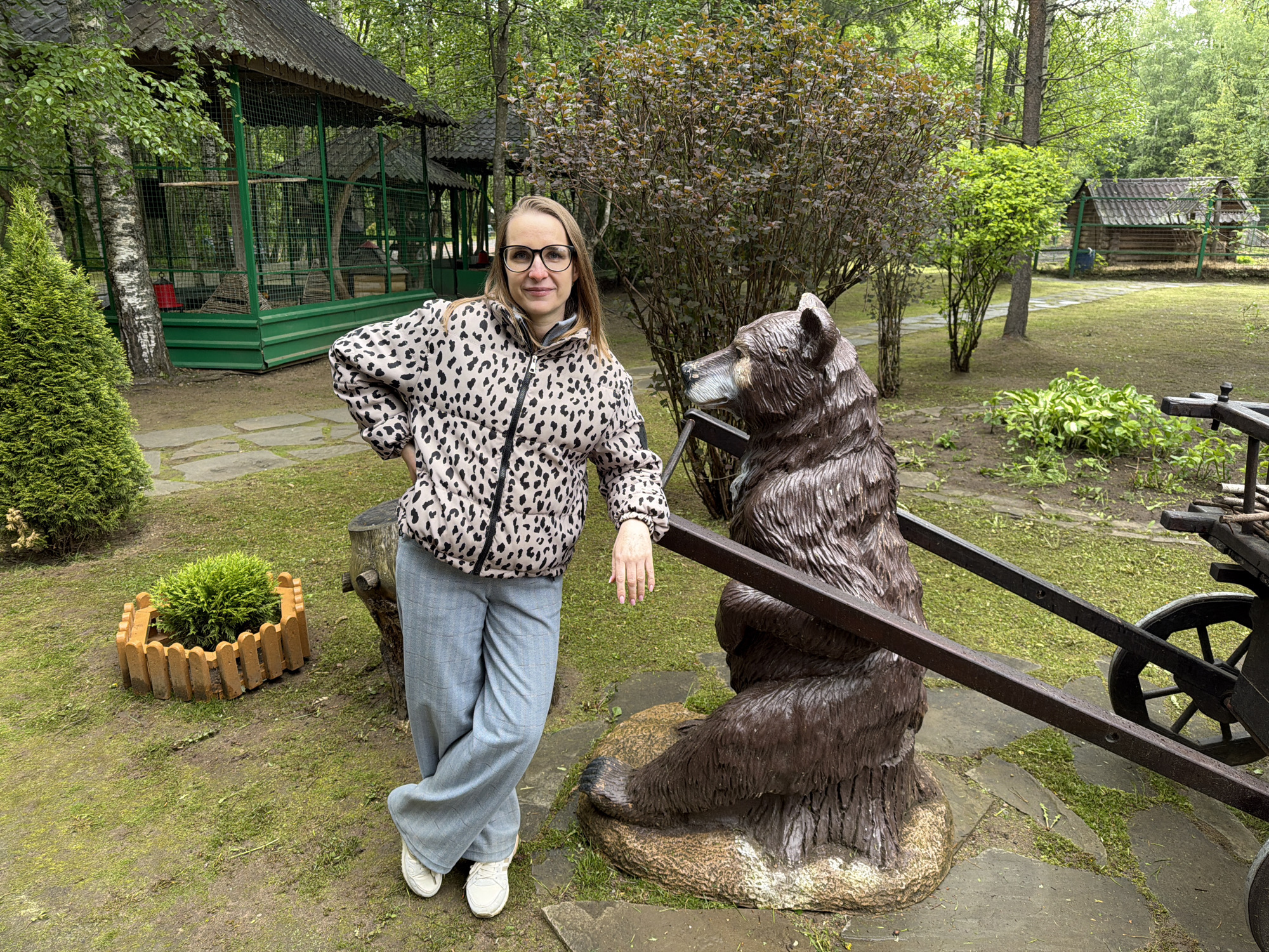 A woman in glasses, a patterned jacket, and gray pants poses beside a wooden bear statue in a park setting, likely in Zaplav'e, Russia. The woman stands with one hand on her hip and the other resting on the statue's carriage attachment. The park features green grass, trees, a wooden gazebo-like structure with cages, and a small log cabin visible in the background. A round plant bed sits near the gazebo. A carved wooden bear statue is the main object the woman is standing with. The ground appears wet, suggesting recent rain.