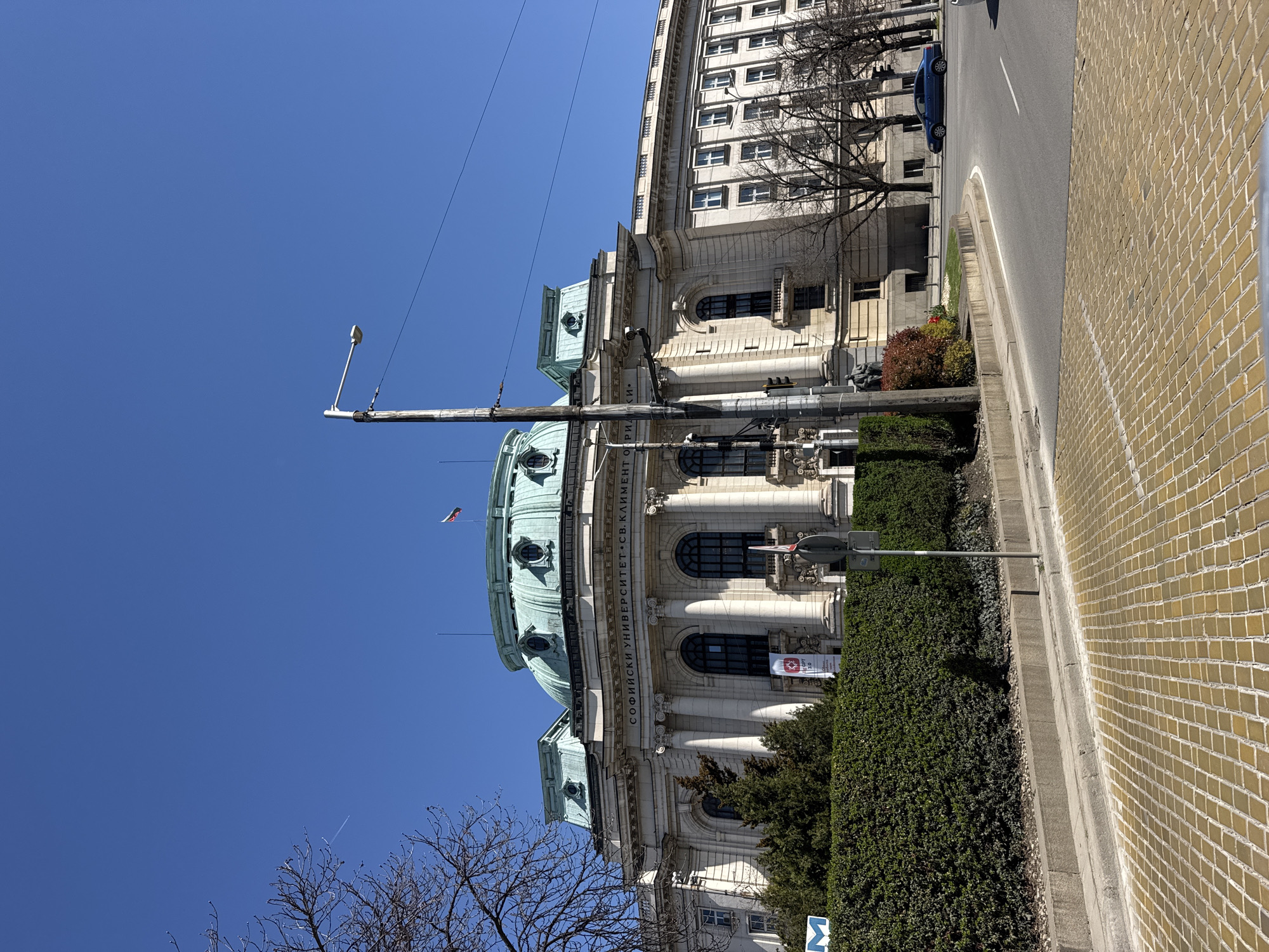 The image depicts the building of Sofia University "St. Kliment Ohridski" in Sofia, Bulgaria. The architectural style of the building is classical, featuring a prominent dome and façade adorned with columns. The building's façade is white, and the dome is a greenish color. In front of the building is a well-maintained green hedge. Alongside the hedge, there is a paved walkway composed of yellow bricks. A street runs parallel to the building, with a blue car parked on the side. In the foreground, a lamppost stands tall against a clear blue sky. A building with many windows is to the right of the university building.