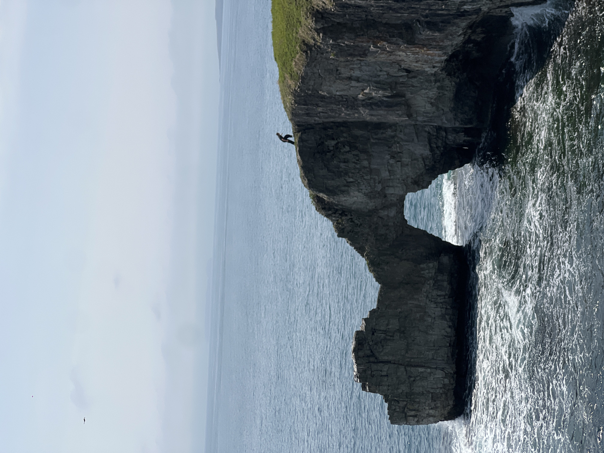 The image captures a coastal scene featuring a dramatic rock formation with a natural archway carved by the sea. The rock is dark grey and textured. A small figure stands on the top of the rock. The sea is visible through the archway, appearing light blue and calm in the distance, with darker, more turbulent water closer to the rock face. The coastline is characterized by a grassy area atop the cliffs. The location is the Bruce Peninsula near Slavyanka, Russia.