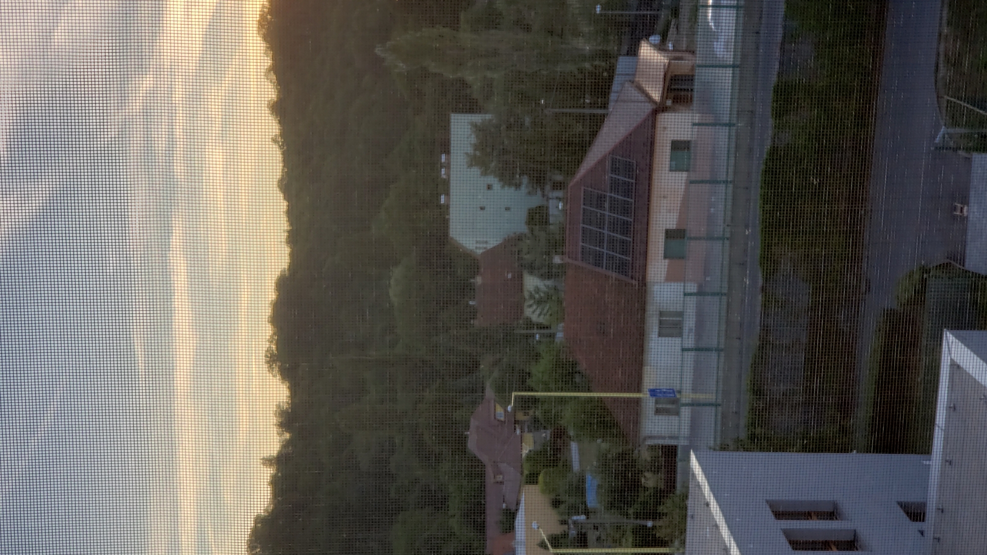 This is a photograph taken from a window with a screen. The scene depicts a residential area with houses, some of which have solar panels on their roofs. A hill covered in trees rises behind the houses. In the distance, a larger, light-colored building can be seen. The sky above is partly cloudy with warm tones suggesting either sunrise or sunset. The image appears to be of a street in the city of Prešov, Slovakia, likely in a suburban or semi-rural area.