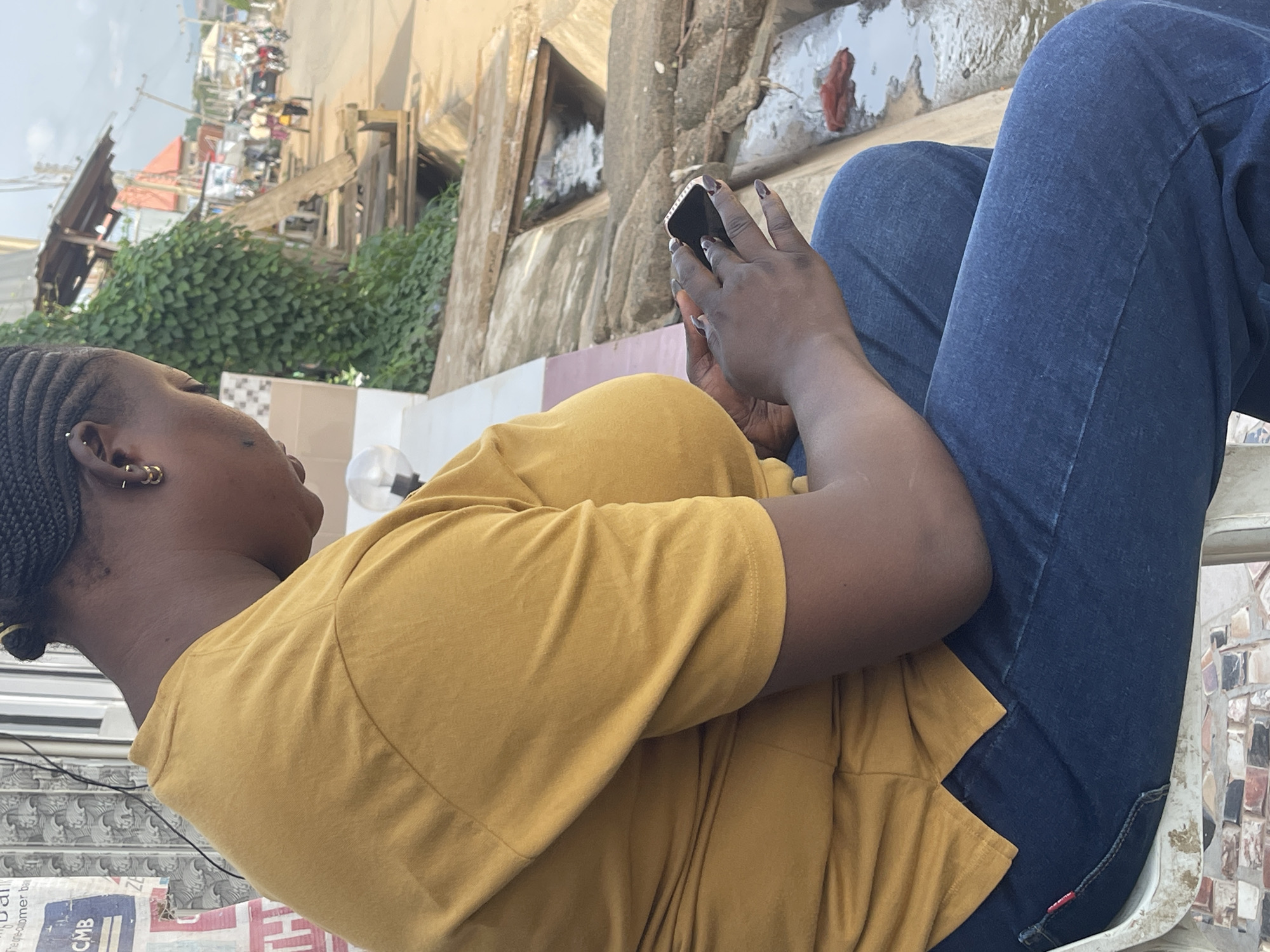 A medium shot captures a Black woman, likely in her late 20s to early 30s, seated outdoors and looking down at her mobile phone. She has braided hair and is wearing a mustard yellow t-shirt and dark blue jeans. Her right hand is holding the phone, and her left hand is resting on her lap. The background shows a busy street scene, suggesting an urban environment. A concrete drainage channel with flowing water is visible to her right, and beyond that, a street with various stalls, vehicles, and pedestrians can be seen. The setting appears to be a roadside area in Abuja, Nigeria, characterized by its bustling atmosphere and typical urban infrastructure.