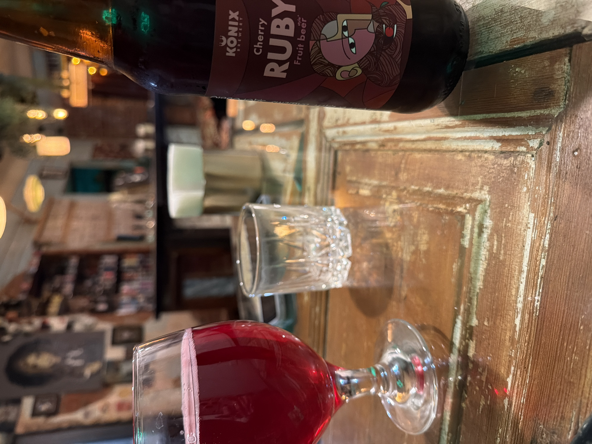 The image depicts a bottle of "Konix Brewery Cherry Ruby Fruit Beer" on a distressed wooden surface in what appears to be a restaurant or bar setting. A wine glass filled with a red beverage sits below, and a small, empty glass is positioned between the bottle and wine glass.
In the blurred background, shelves are stocked with various items, possibly bottles and merchandise. There is a dimly lit wall with framed pictures. The ambient lighting is warm, creating a relaxed atmosphere. The location can not be verified.