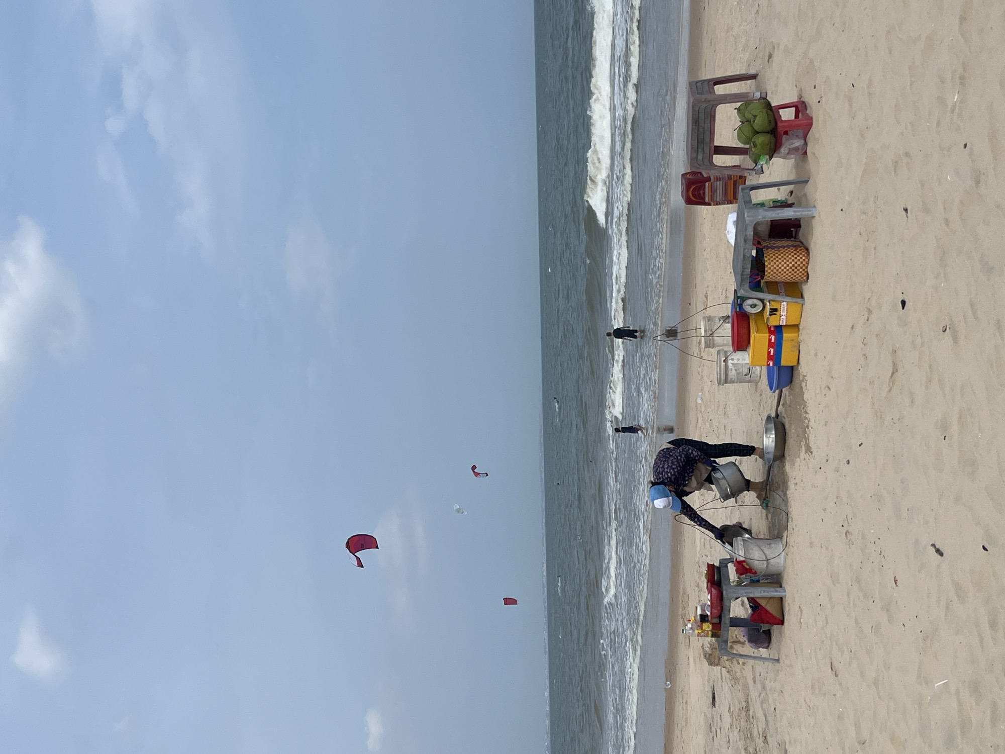 The image depicts a beach scene in Mũi Né, Vietnam, with the ocean in the background and sand in the foreground. Several kites are visible in the sky. On the beach, a person wearing a blue and white cap and a patterned shirt is tending to a small stand. The stand has a table laden with various items, including bottles, bowls, and what appear to be food containers. A metal bucket and a larger metal basin are also present near the person. To the right of this stand, another table displays a stack of coconuts on a small red stool. In the distance, a few individuals are seen in the water. The overall atmosphere is that of a typical beach day with recreational activities and a vendor selling refreshments.