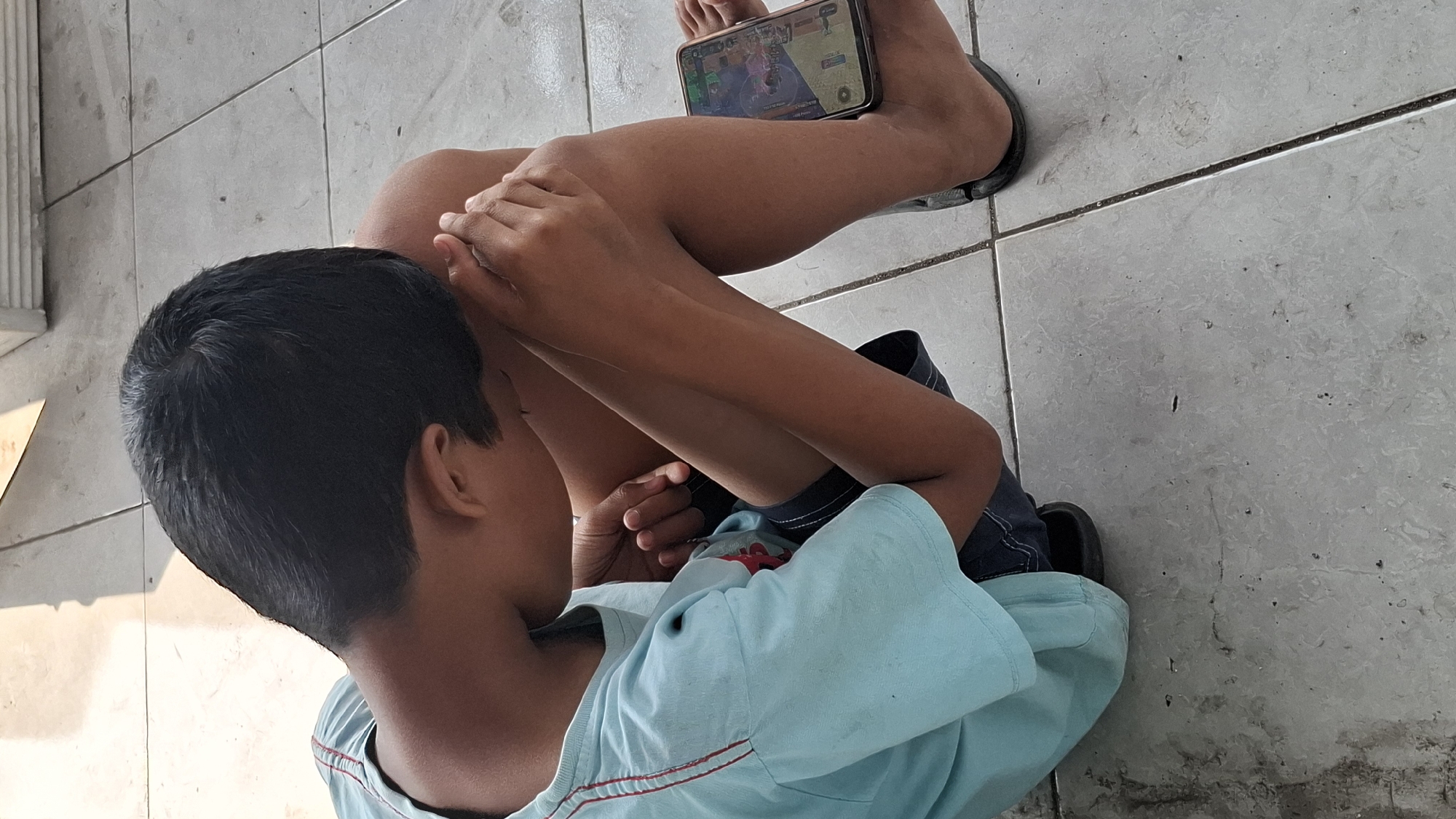A young boy is sitting on a tiled floor, hunched over with his head resting on his knees. He is wearing a light blue t-shirt with red trim and dark blue shorts. A smartphone is placed on his right leg, displaying a game interface. His left foot is bare, and his right foot is partially visible wearing a black sandal. The floor appears to be an outdoor patio or walkway, with distinct tile grout lines. Sunlight casts a shadow on the floor from an unseen object to the left. The overall scene suggests a moment of quiet engagement with a mobile game in a domestic outdoor setting in Cilaku, Indonesia.