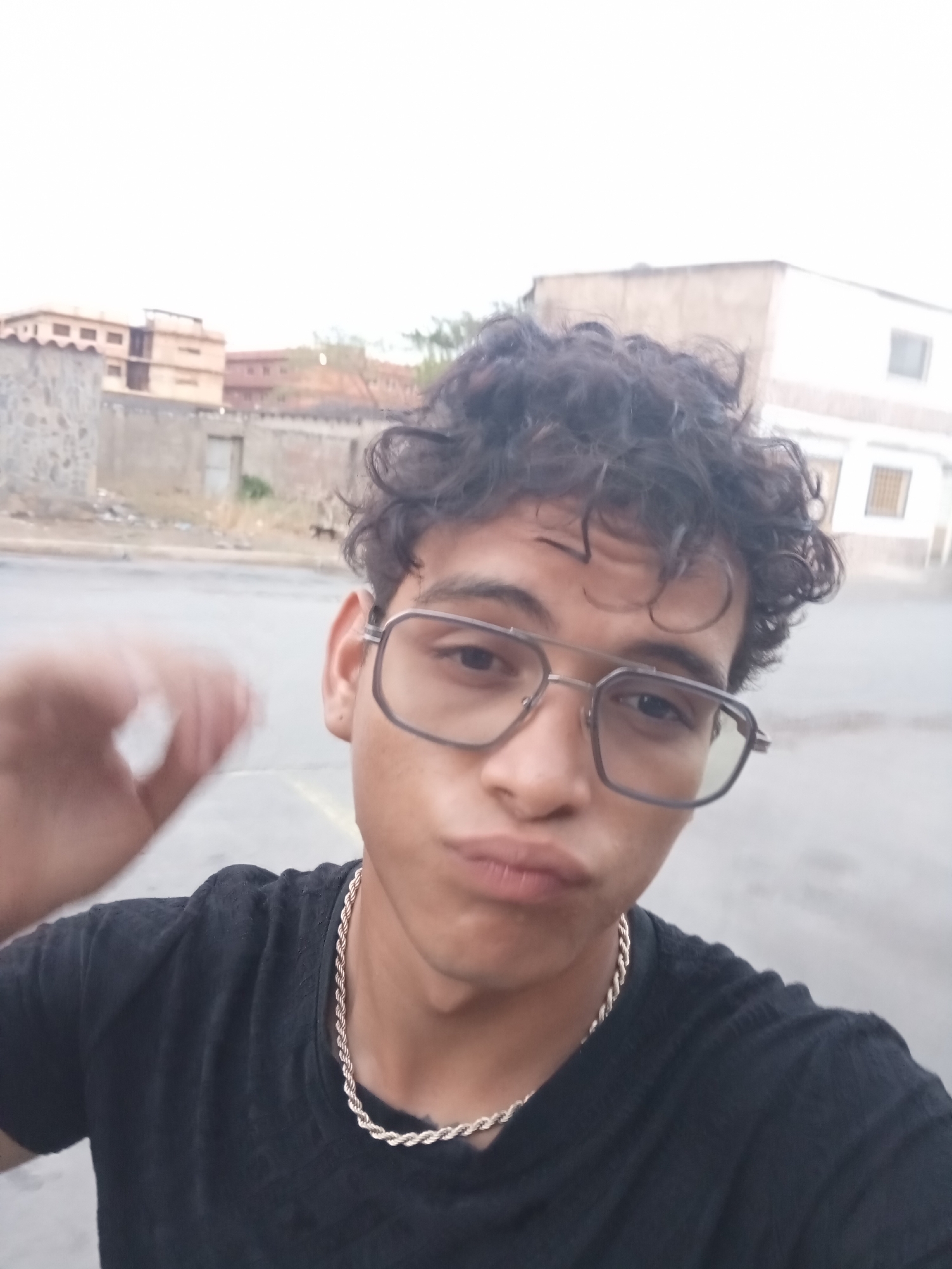 A young man with dark, curly hair and glasses is featured in a close-up, slightly off-center selfie. He is wearing a black t-shirt and a gold-colored chain necklace. His lips are pursed in a "pout" expression, and his eyes are looking directly at the camera through his rectangular, metal-framed glasses. His left hand, blurred due to motion or proximity to the camera, is raised on the left side of the frame, possibly mid-gesture or adjusting something.
The background shows an outdoor, somewhat rustic urban or semi-urban setting, consistent with Juan Griego, Venezuela. Distant buildings, including multi-story structures with terracotta-colored roofs and lighter-colored facades, are visible. Some low stone walls and sparse, dry-looking vegetation can also be seen. The ground appears to be a paved area, possibly a street or an open lot. The sky is bright and appears overcast or diffused, indicating daytime, likely late morning or afternoon, without direct harsh sunlight. There is no visible text in the image.