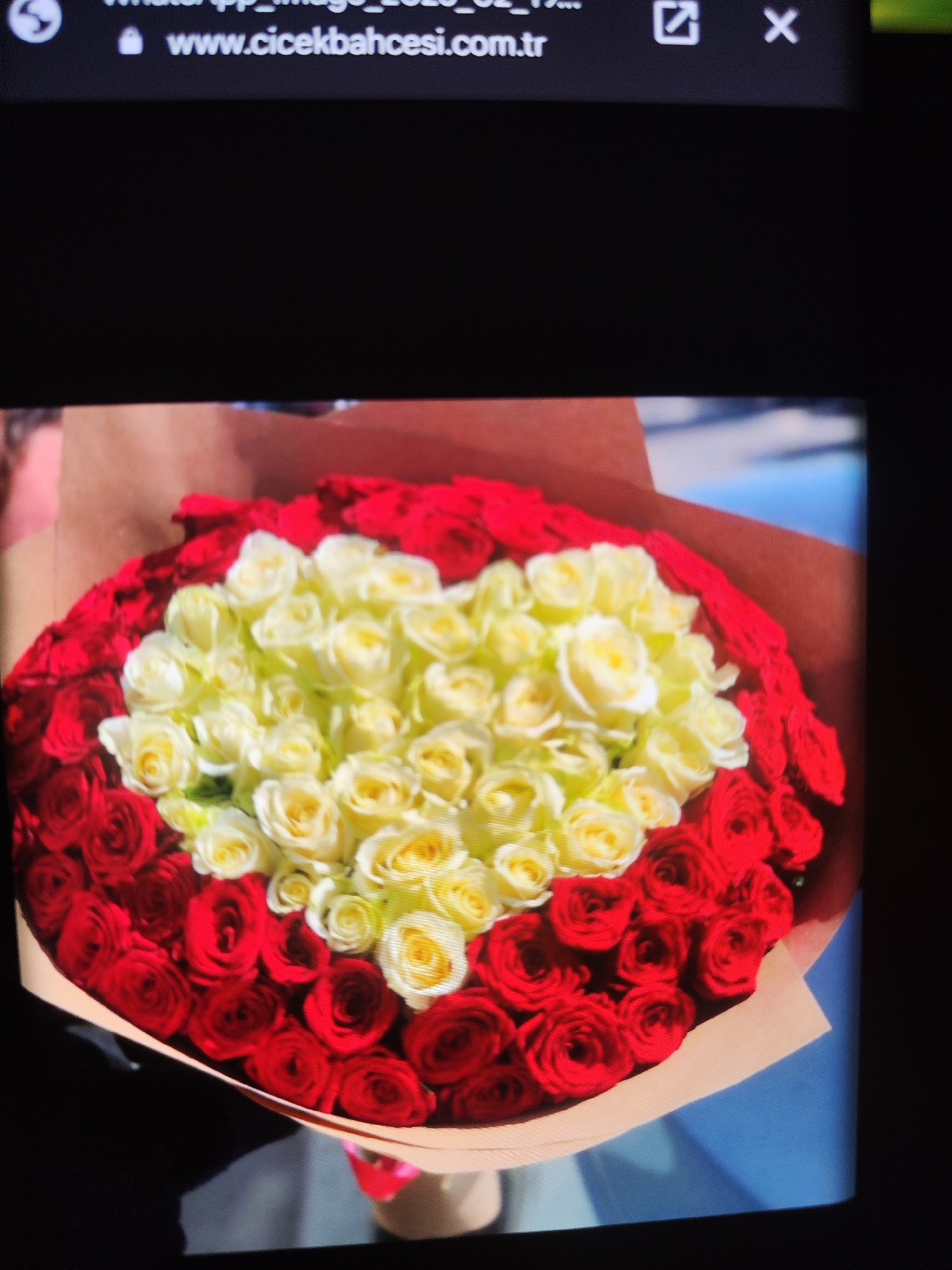 A close-up, overhead shot shows a bouquet of red and yellow roses. The bouquet is wrapped in brown paper and is likely a gift. In the background, a laptop screen displays a web address, "www.cicekbahcesi.com.tr", suggesting the image was taken in front of a computer. There is no discernible presence of the city of Küçükçekmece, Turkey, in this image.